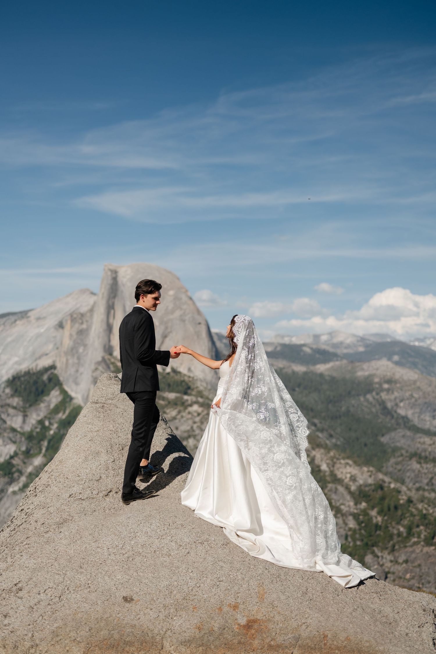 Bride and groom hold hands on a rocky overlook at Yosemite with Half Dome and blue skies behind them.