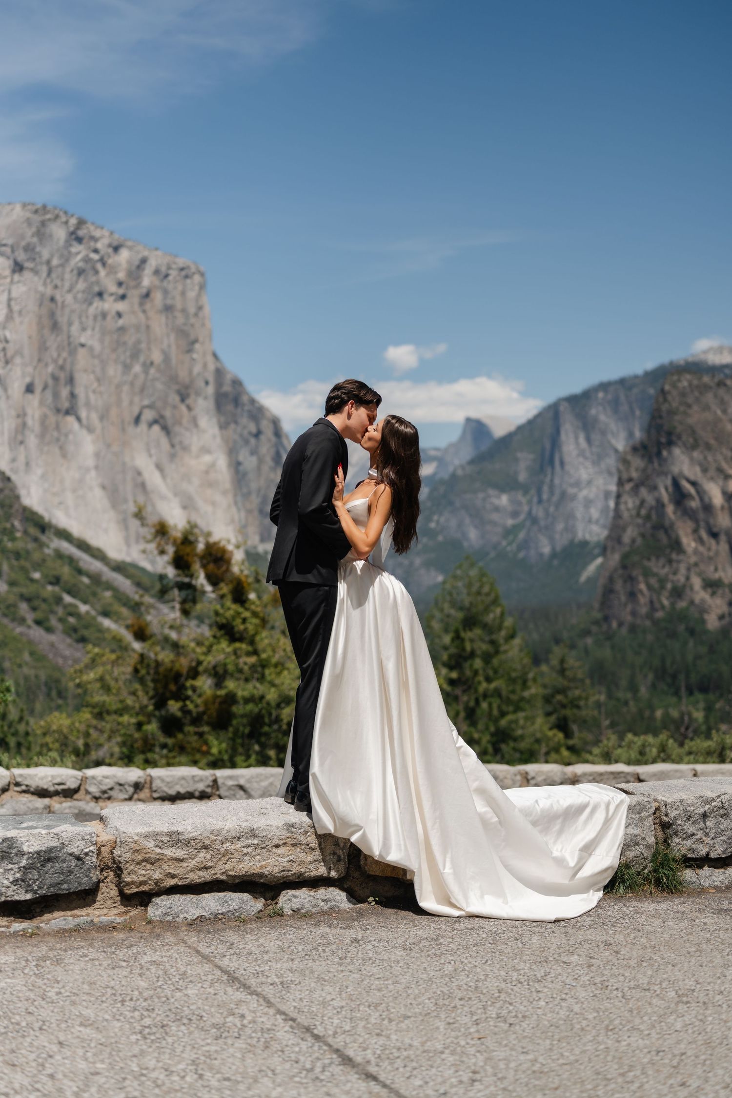 Bride and groom share a romantic kiss at Yosemite Valley overlook with El Capitan and Half Dome in background.