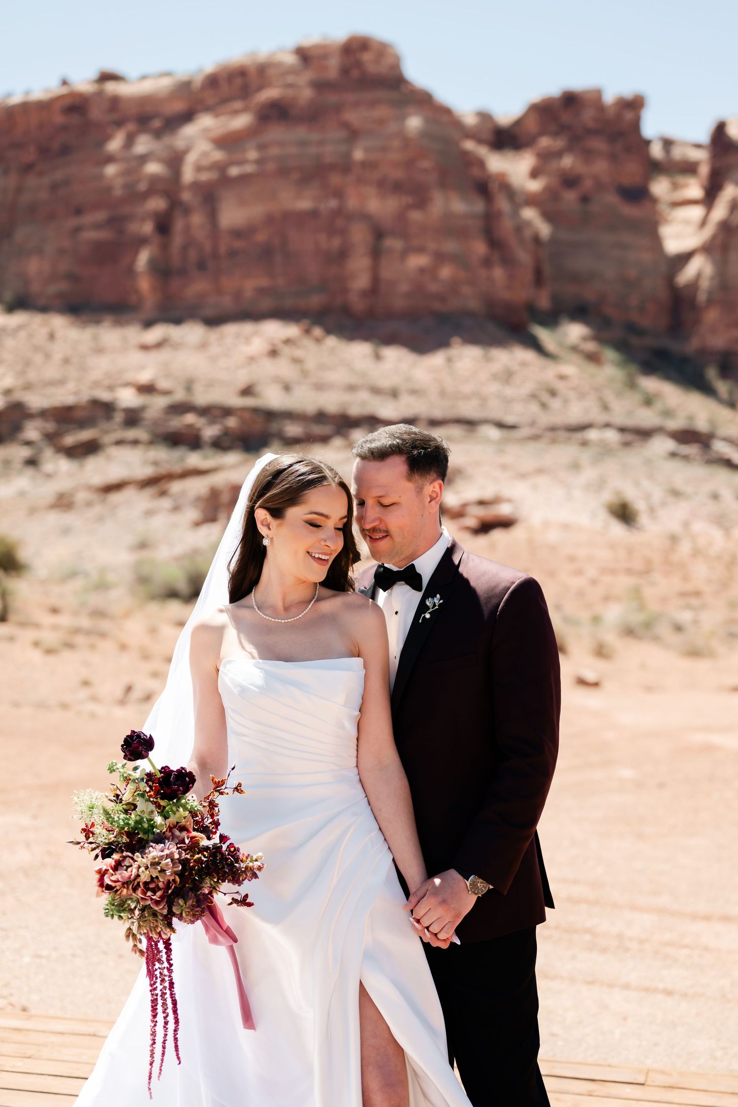 Bride and groom embrace in desert landscape with red rock formations, she holds a dark floral bouquet in white gown.