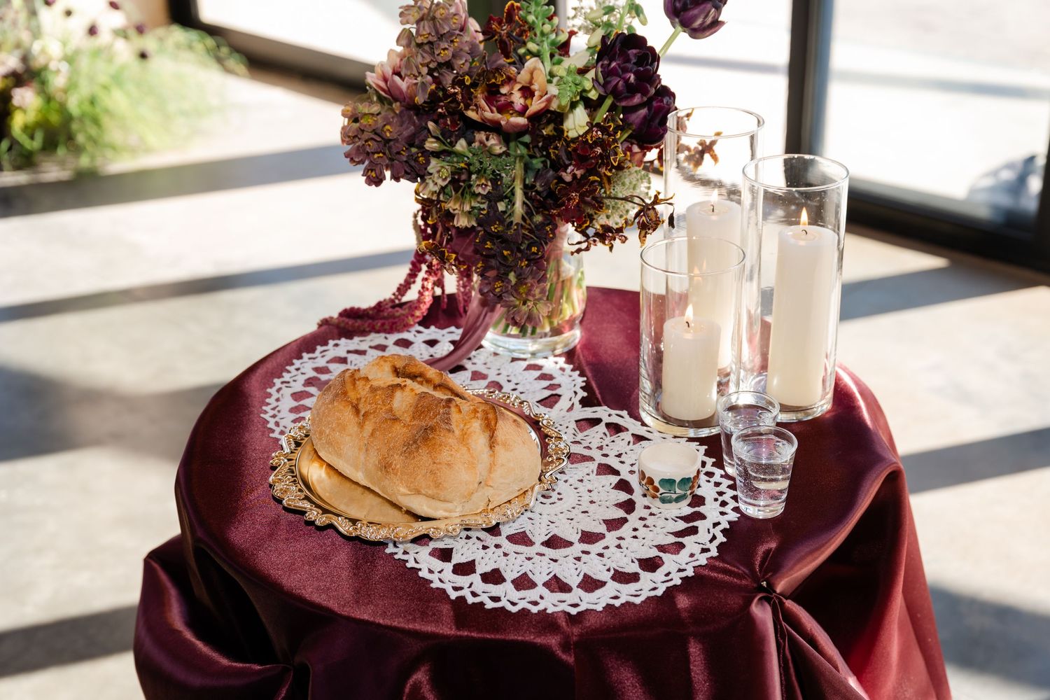 Elegant wedding table with burgundy linen, bread loaves on lace doily, white candles, and purple floral centerpiece.