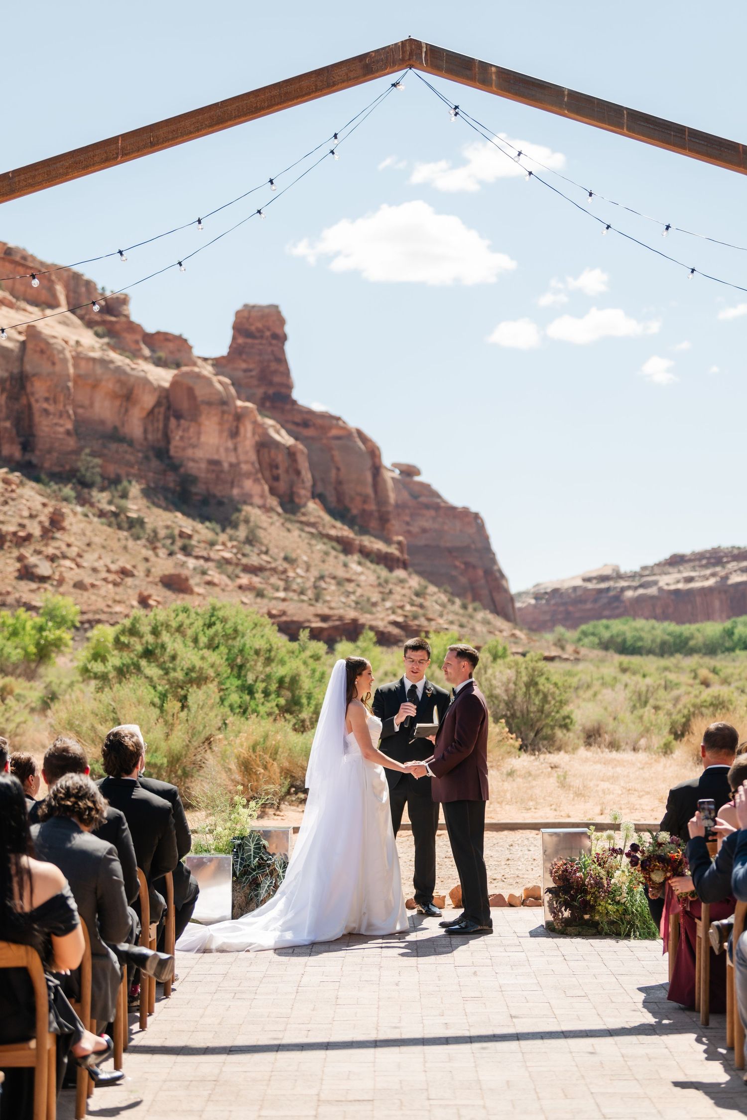 Outdoor wedding ceremony with bride and groom at altar, framed by rustic arch, string lights, and stunning red rock canyon backdrop.
