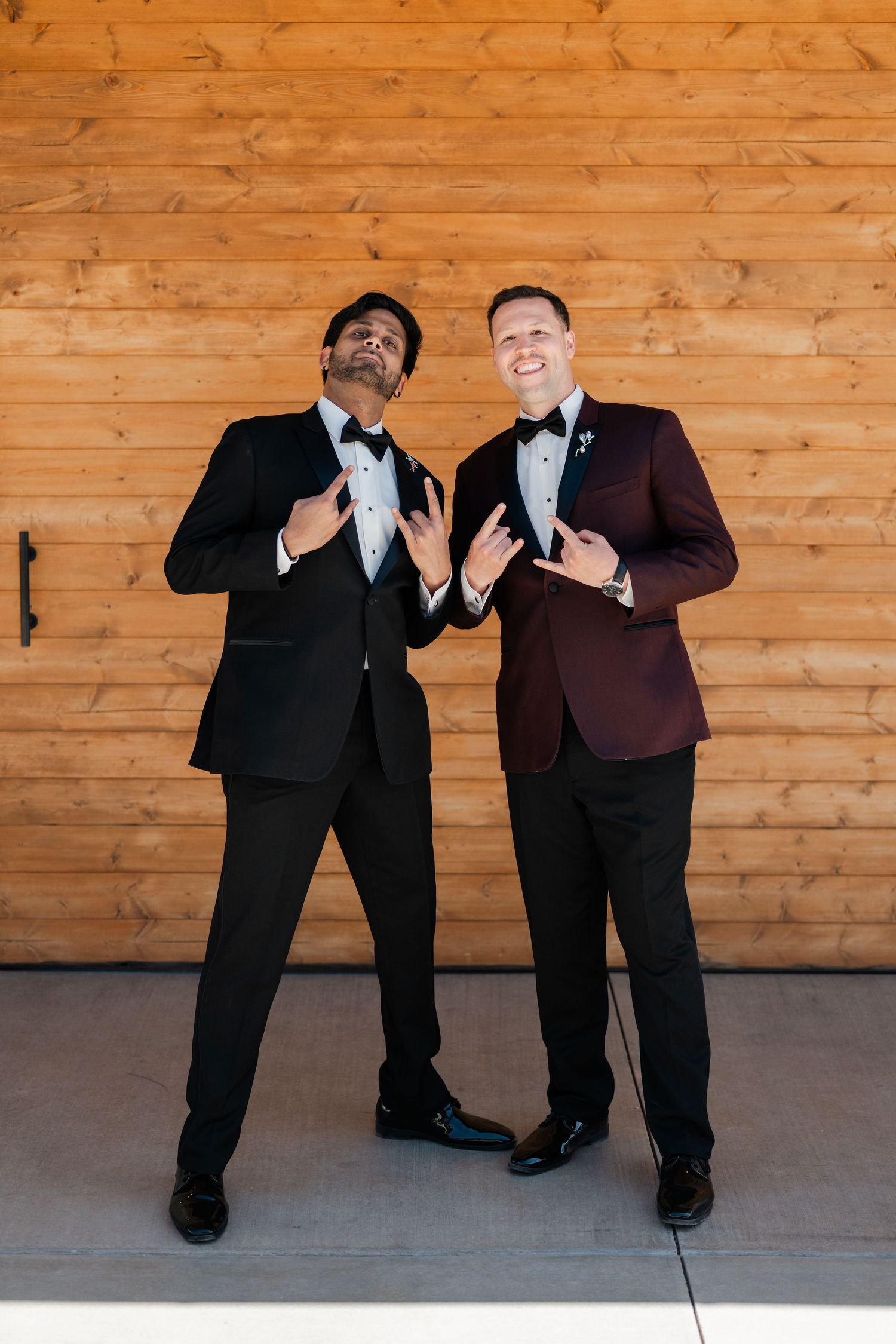 Two groomsmen in tuxedos strike fun poses against wooden barn wall, one in black and one in burgundy jacket.