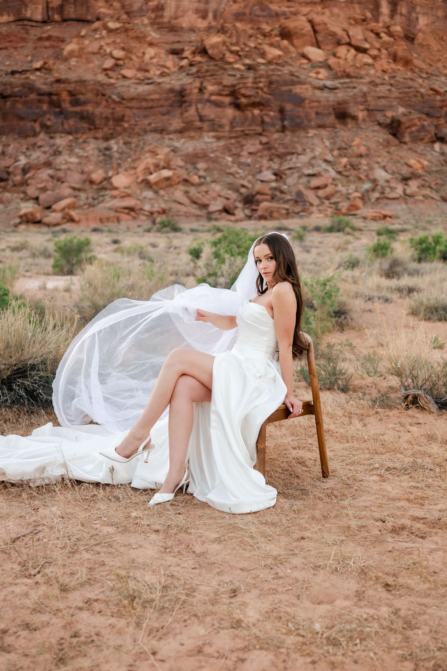 Bride in white strapless gown and veil sits on wooden chair in desert landscape with red rock formations.