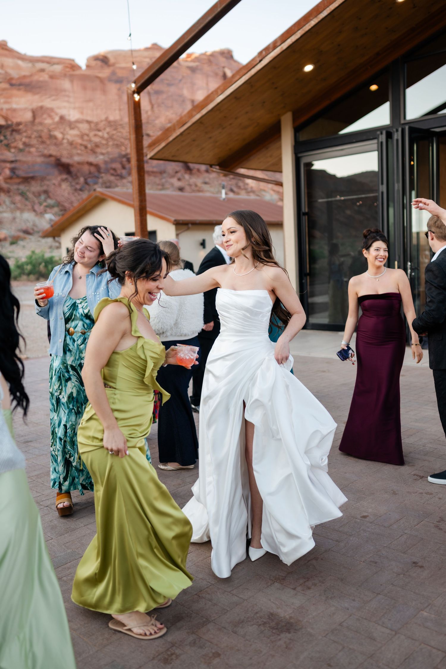 Bride in white gown dances with guest in green dress at outdoor wedding reception near red rock cliffs.