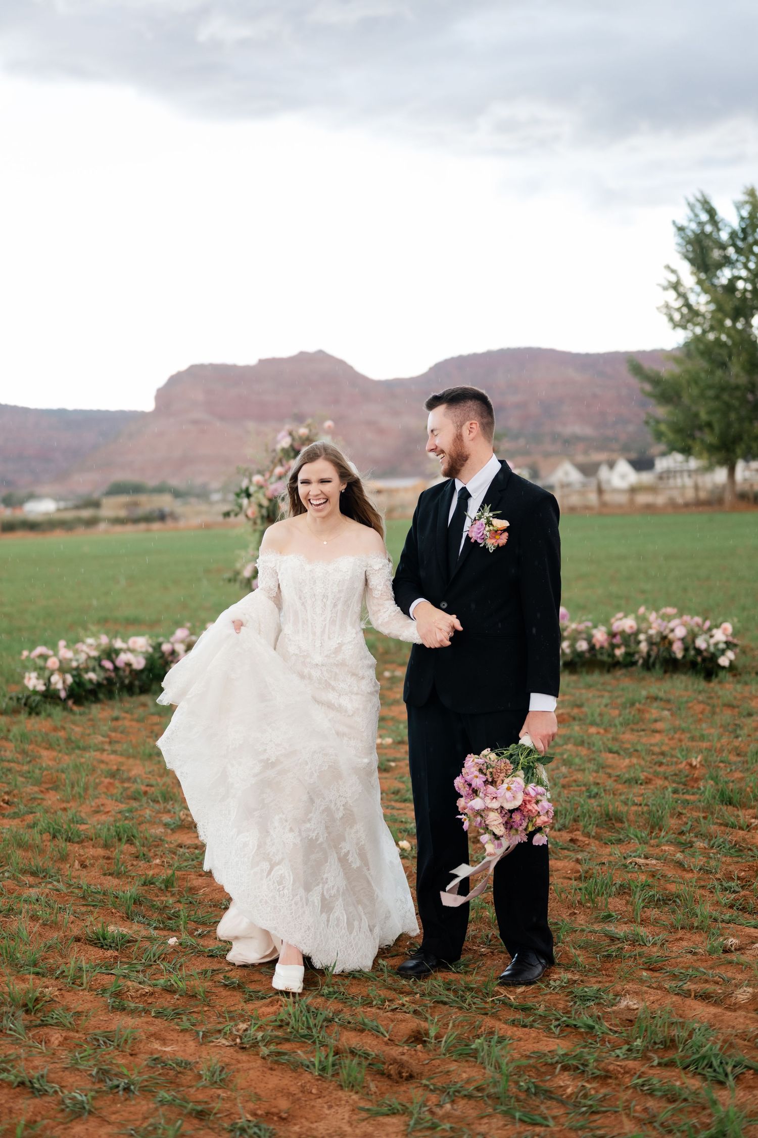 A couple in wedding attire playfully walks through a red desert landscape with mountains. Southern Utah Wedding Photographer.