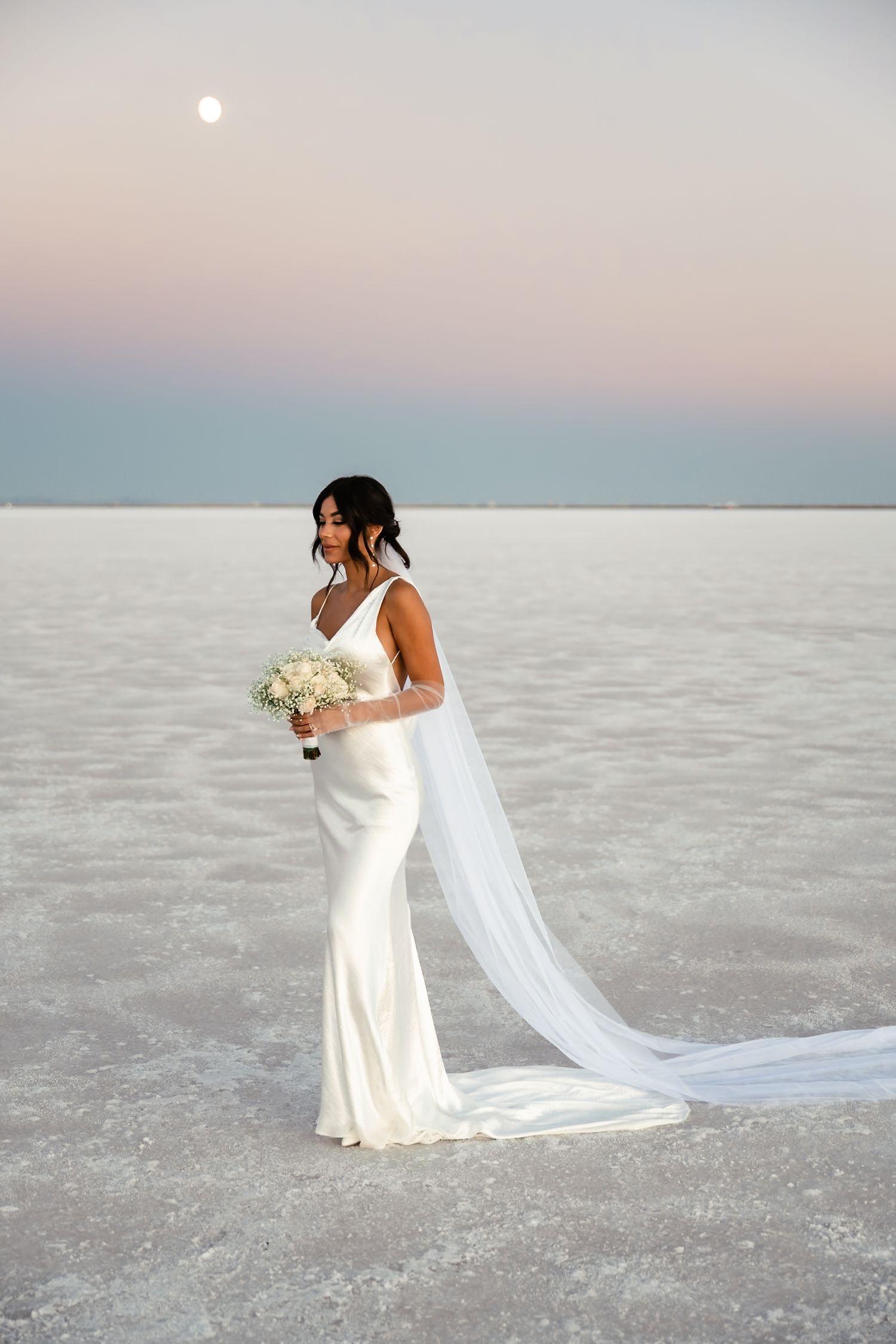 A bride in a fitted white gown with long veil holds a bouquet on salt flats at sunset.