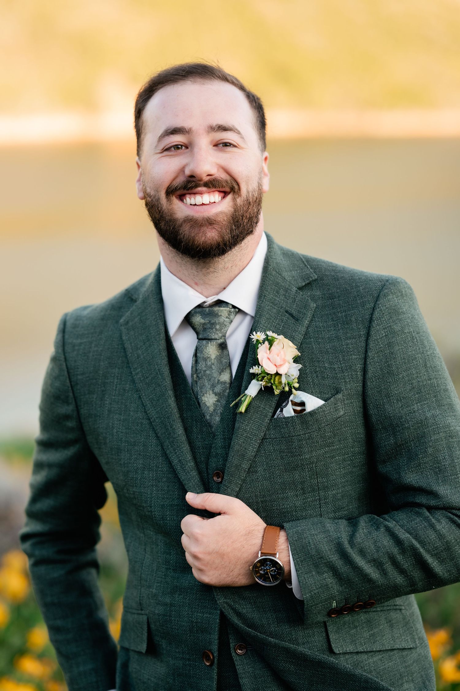 Groom in dark green suit and tie smiles while standing against golden sunset backdrop.