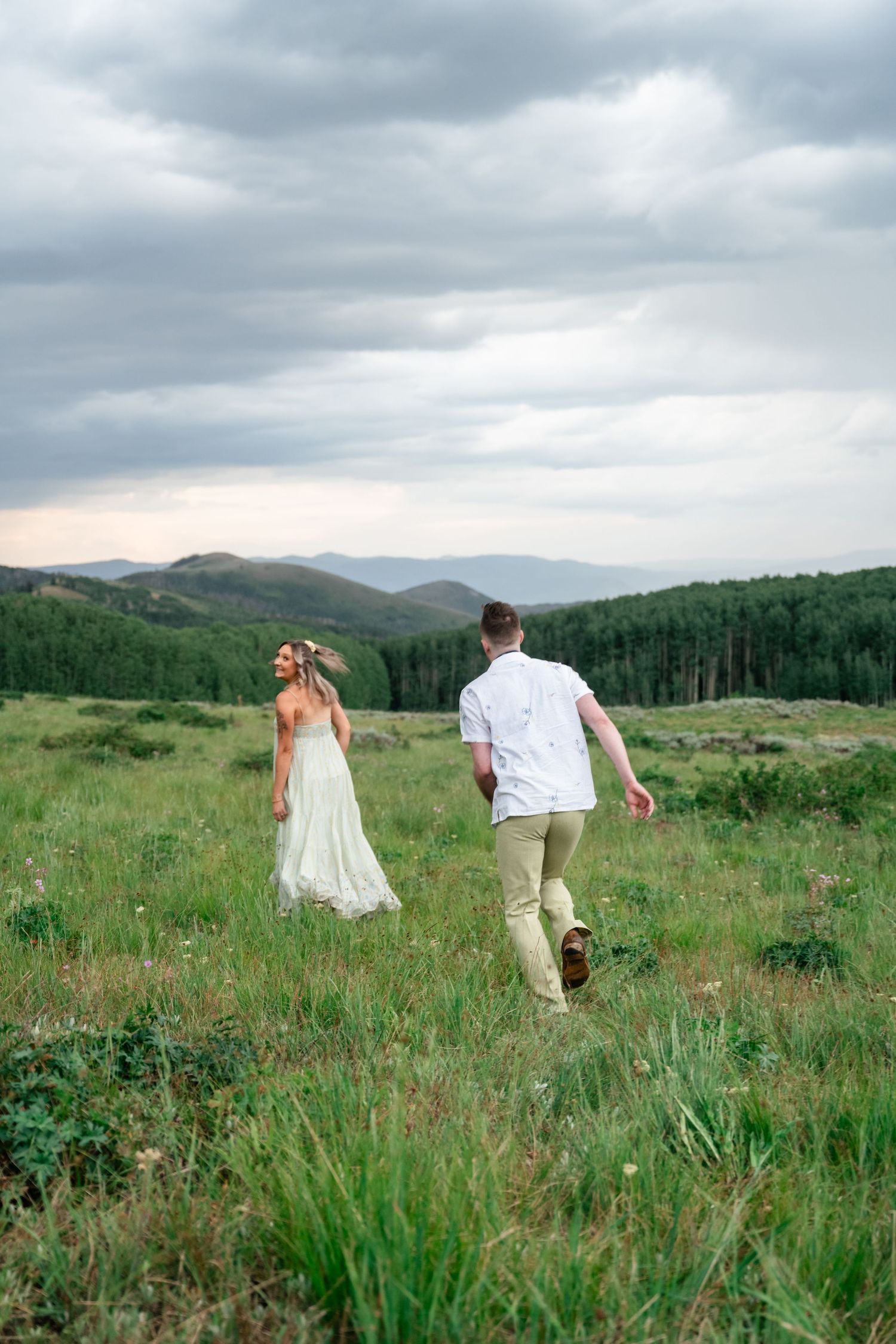 A couple walks through a scenic mountain meadow under dramatic cloudy skies.