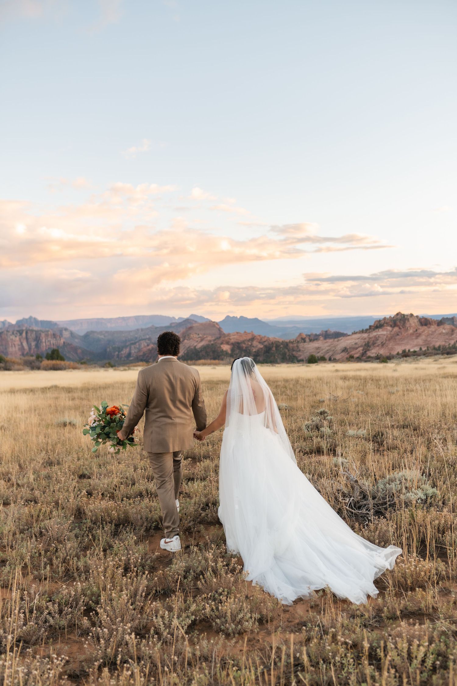 A couple walks hand in hand through a golden field at sunset with mountains in the distance.