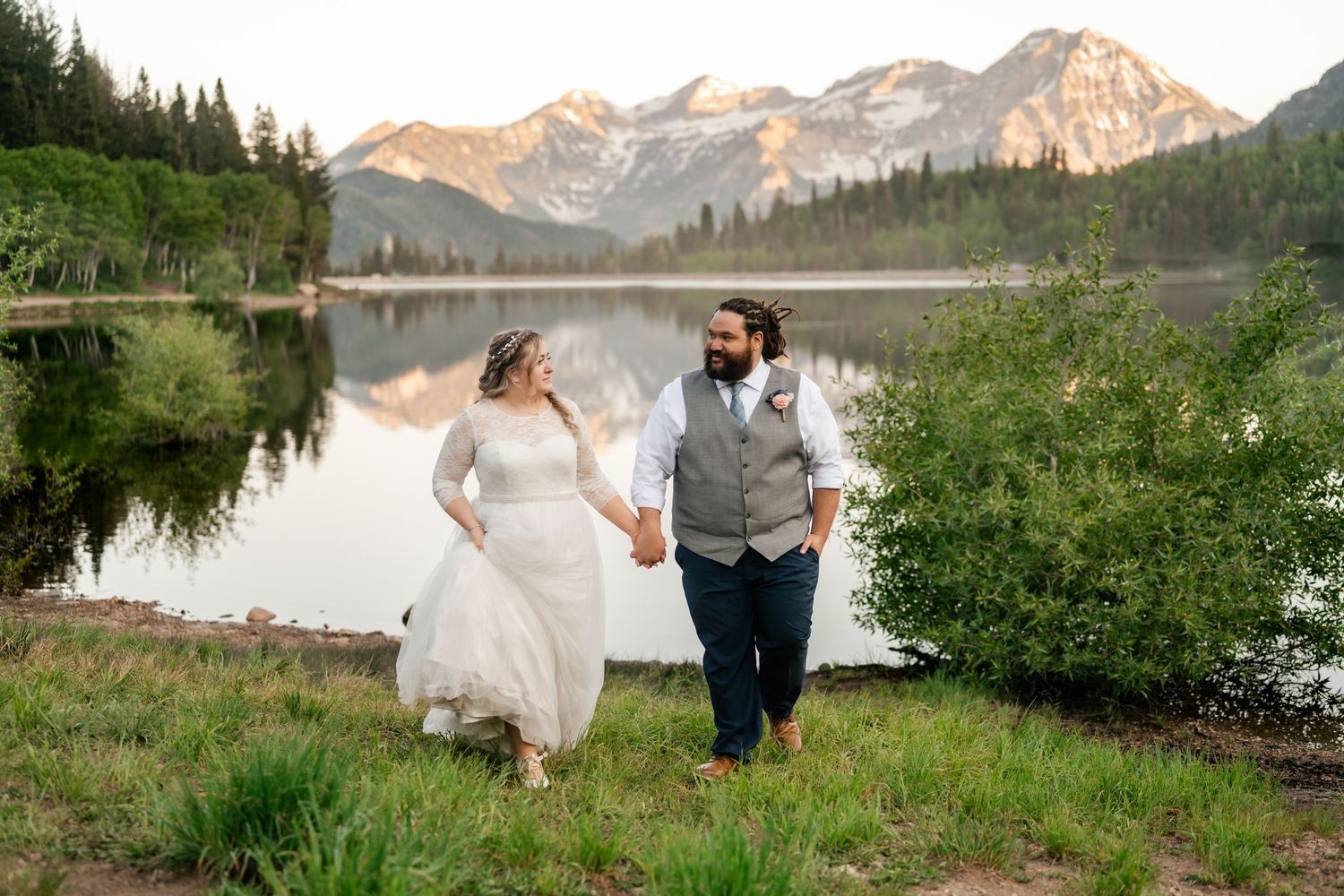 A wedding couple poses against a scenic mountain backdrop near an alpine lake during golden hour. Utah mountain elopement photographer.