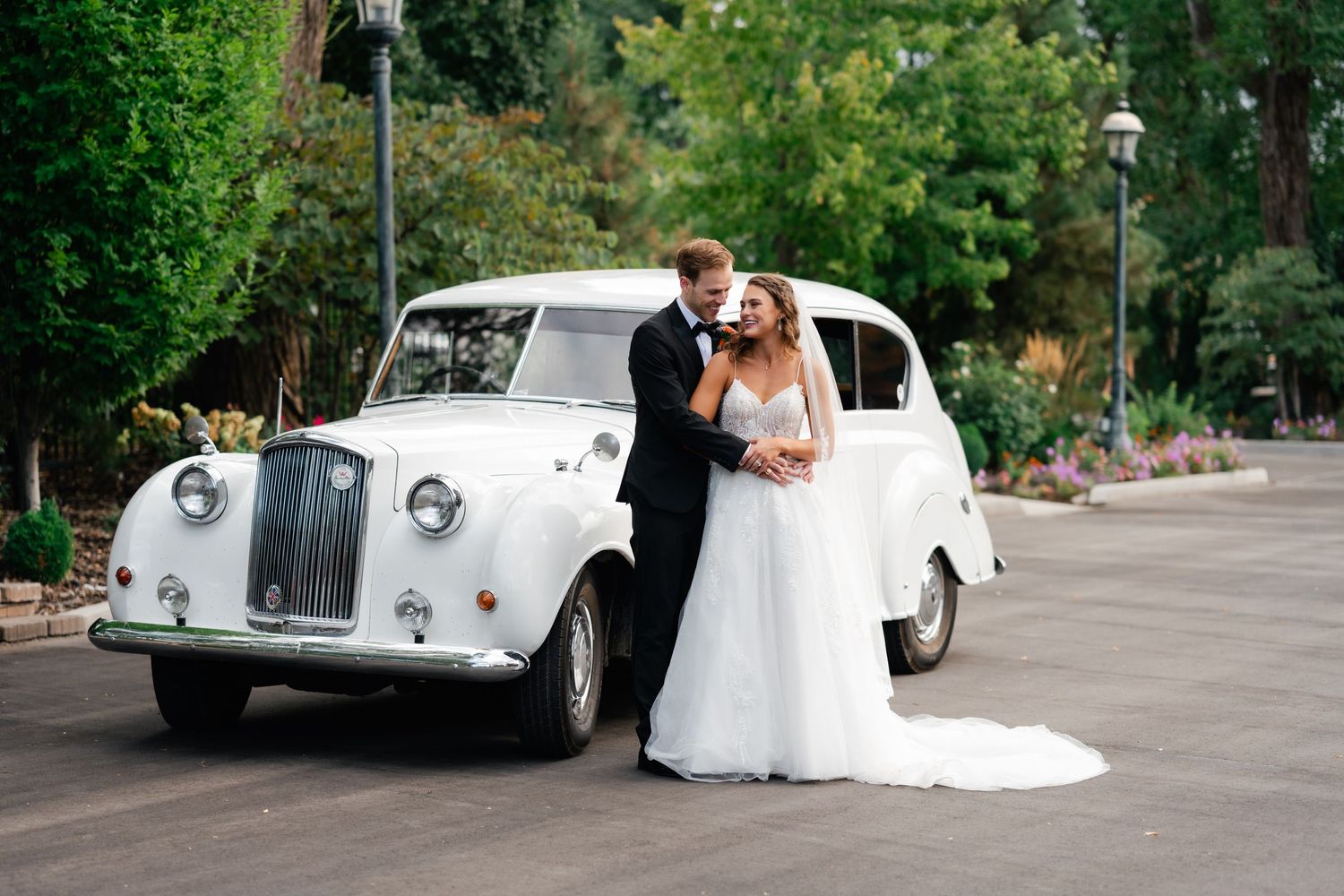 Classic white vintage car parked on tree-lined path with wedding couple posing for romantic photos. Utah luxury wedding photographer.