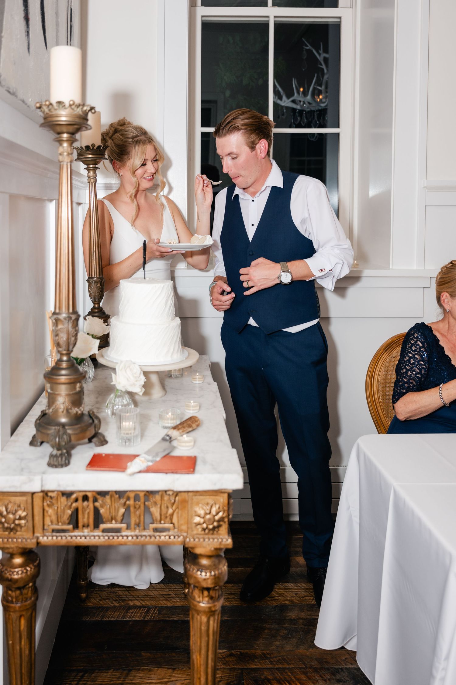Wedding couple and guests share joyful moments while cutting their elegant white cake at an indoor reception.