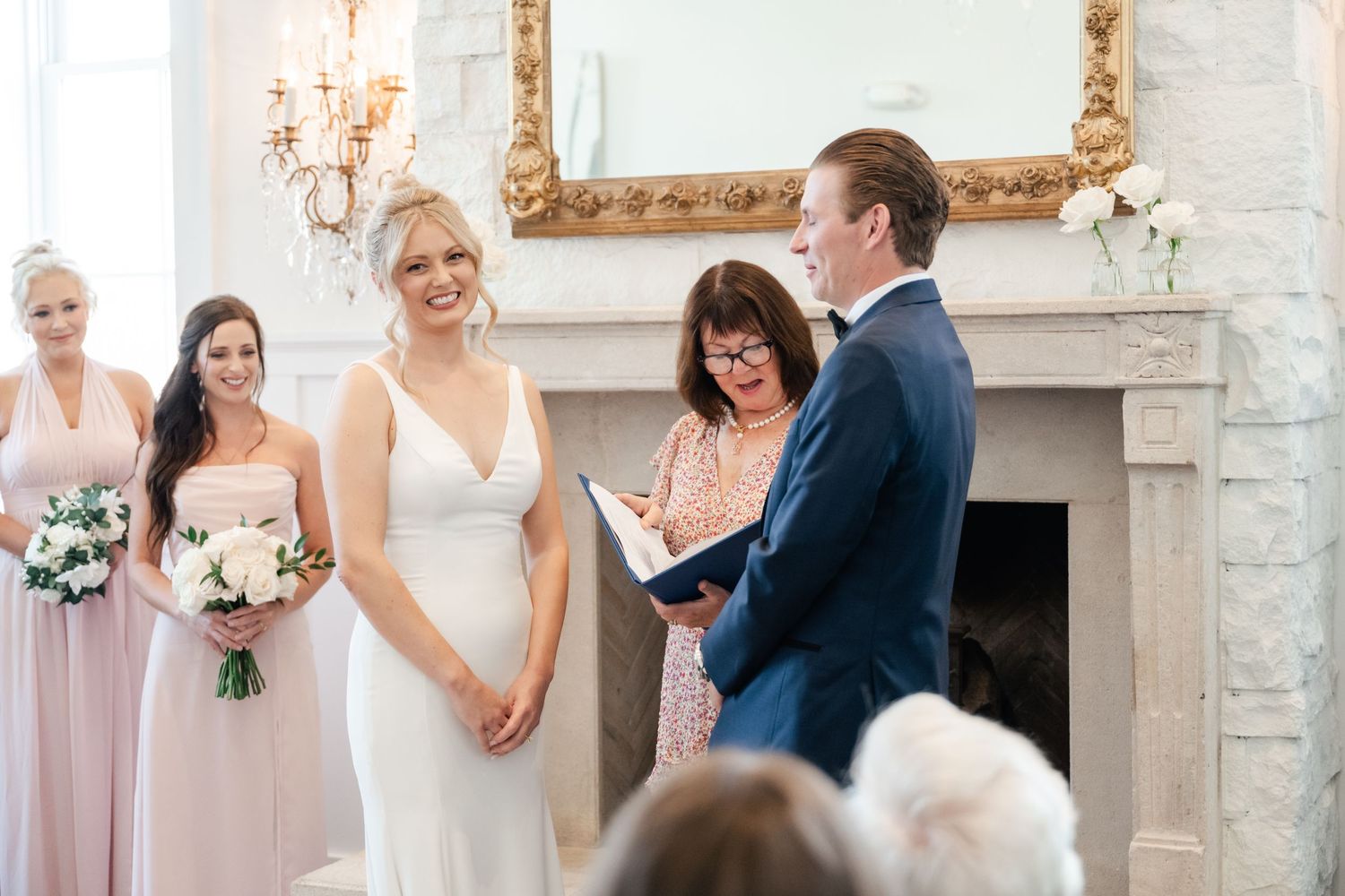 Wedding ceremony takes place in front of an elegant fireplace with ornate gold mirror and white floral bouquets.