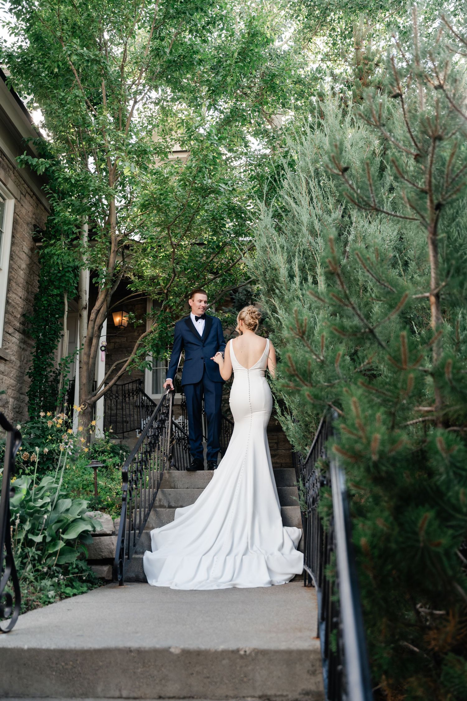 Romantic wedding couple poses in a garden pathway surrounded by lush greenery and trees.