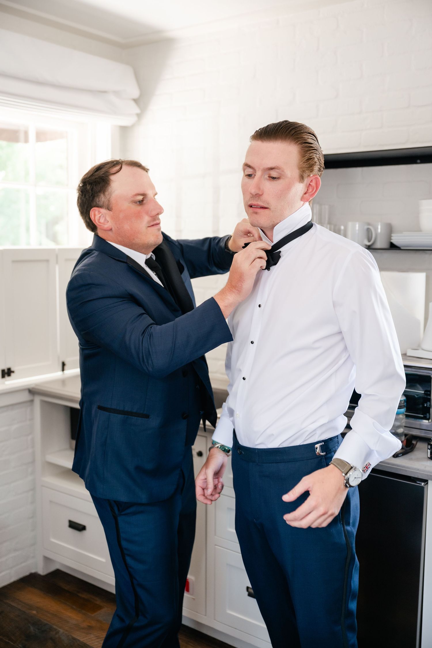 Two people adjusting formal attire in white modern kitchen setting.