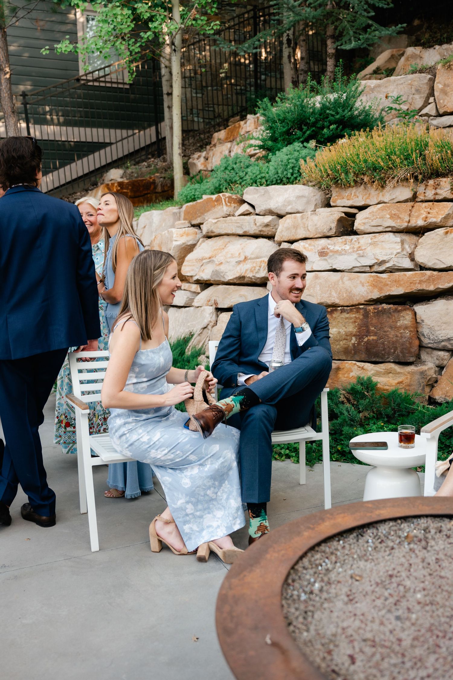 Guests relax in white chairs on a stone patio with landscaped rock wall and greenery in the background.