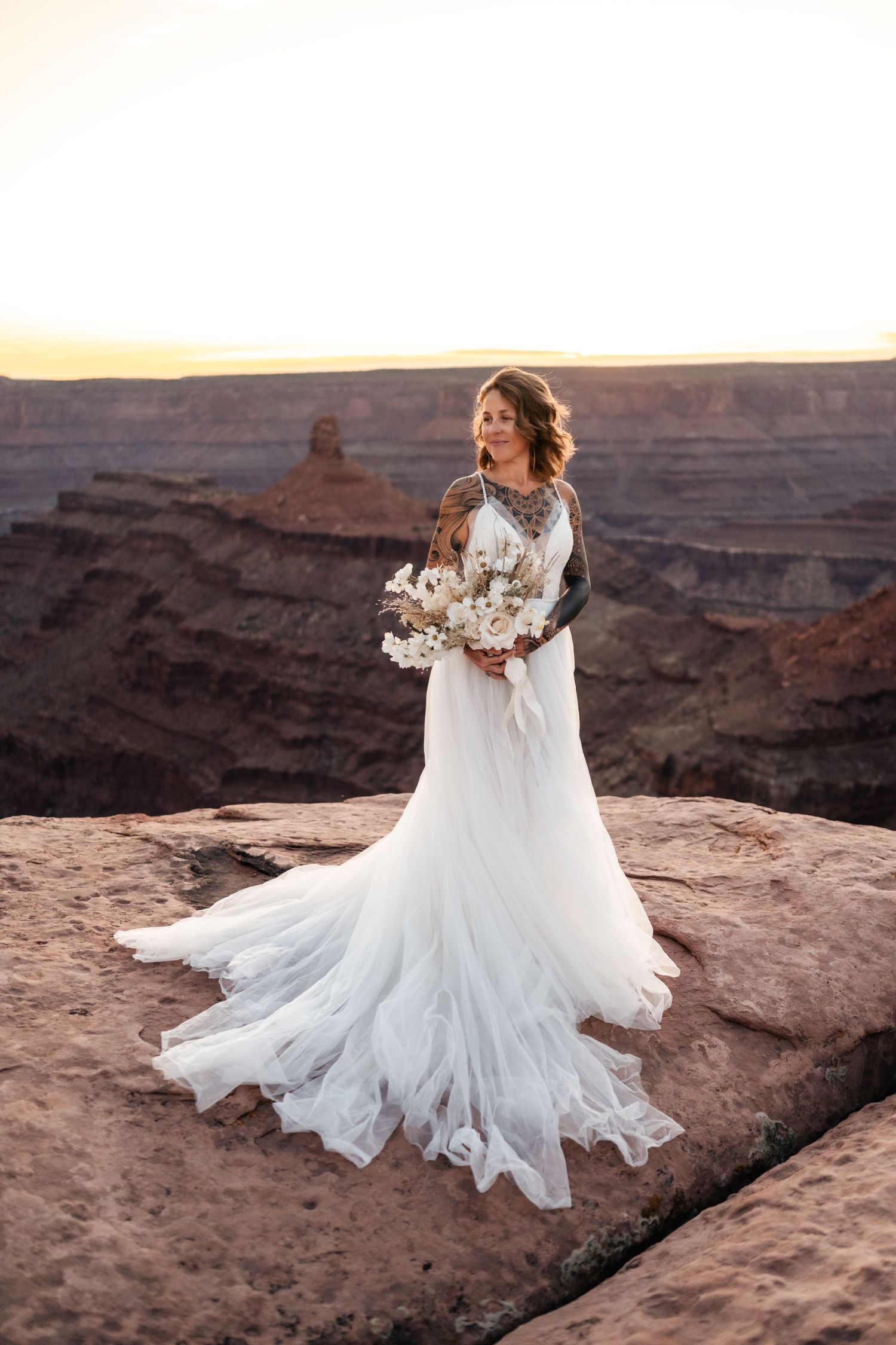 A bride in a flowing white wedding dress on red rock cliffs during sunset at a desert canyon overlook. Dead Horse State Park Photographer.