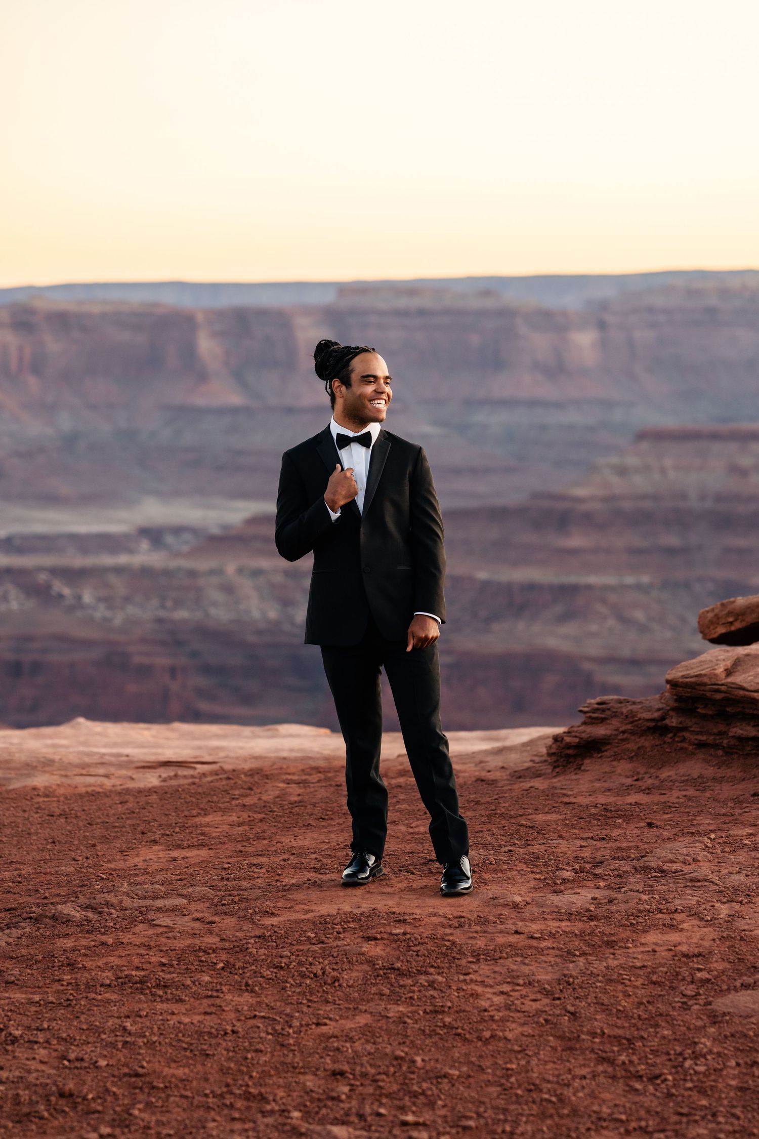 A person in a formal black suit walks across red desert terrain with canyon cliffs in the background. Dead Horse State Park Photographer.