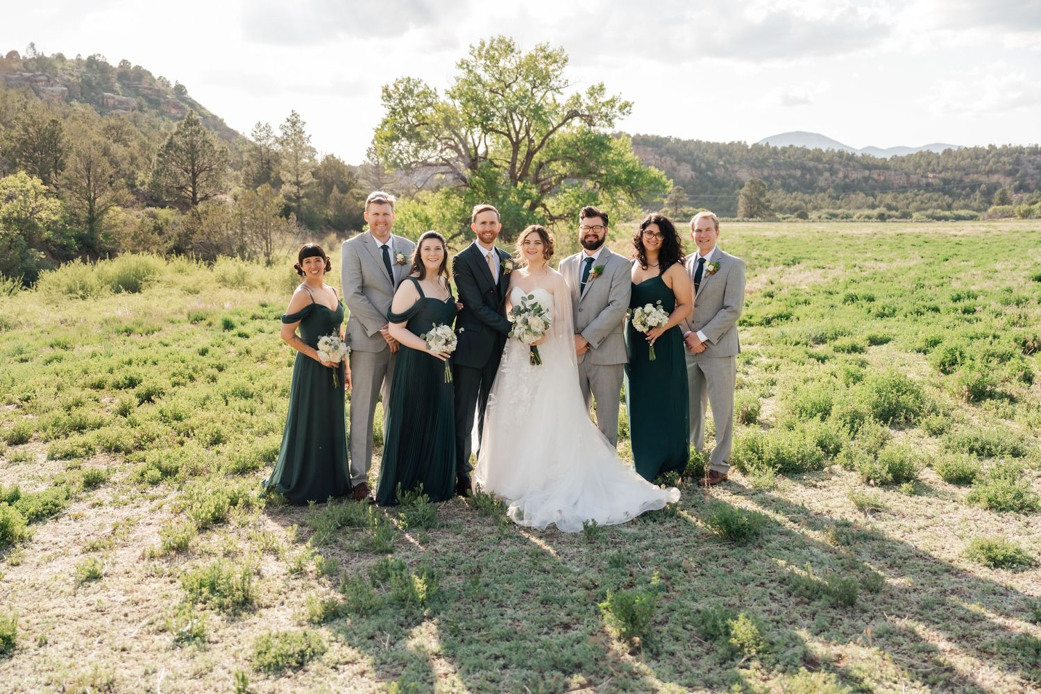 Wedding group photo outdoors with bridal party in dark green dresses and gray suits against mountain landscape.