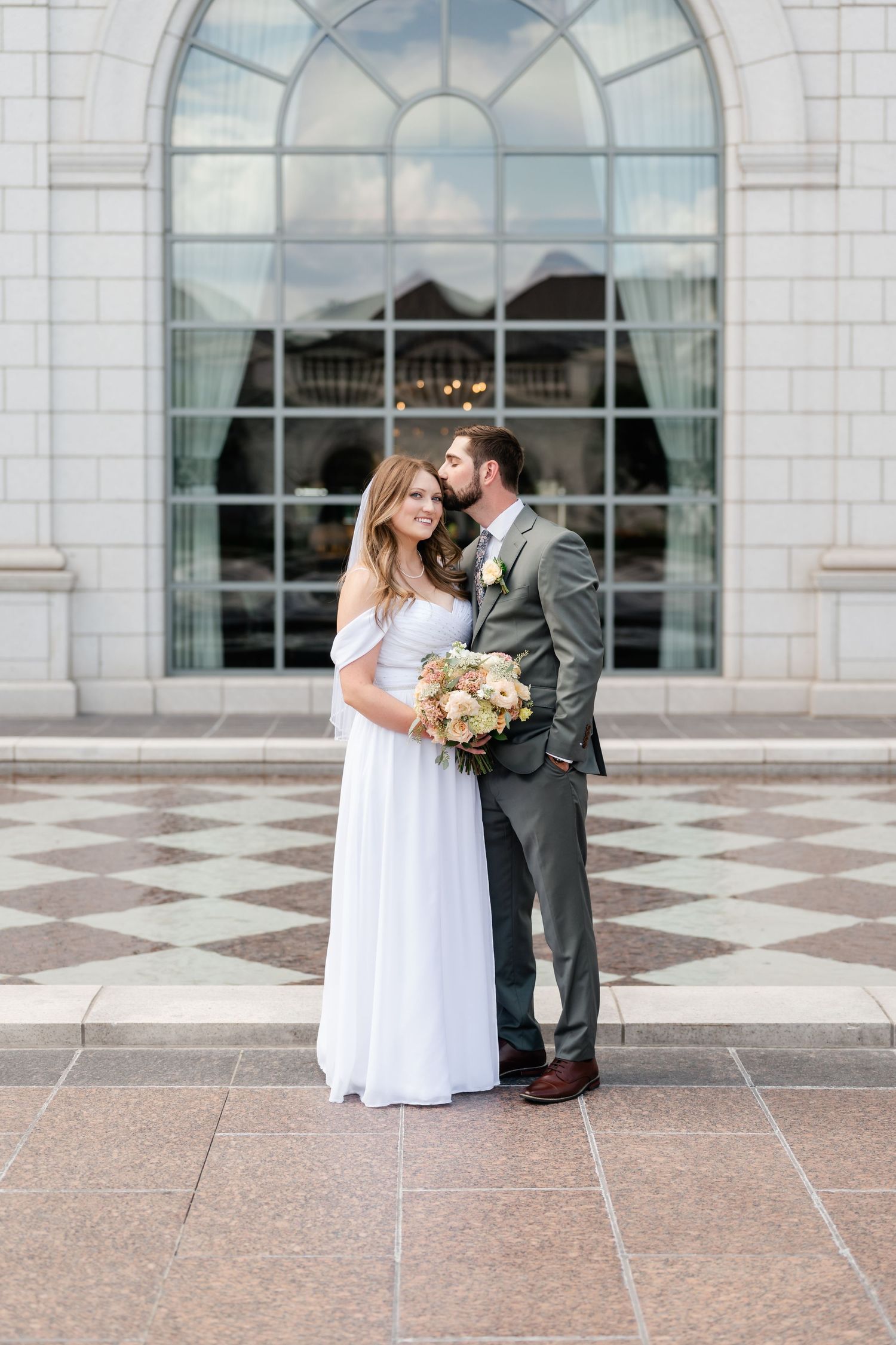 Wedding couple poses in front of a grand building with arched windows and checkered stone floor.