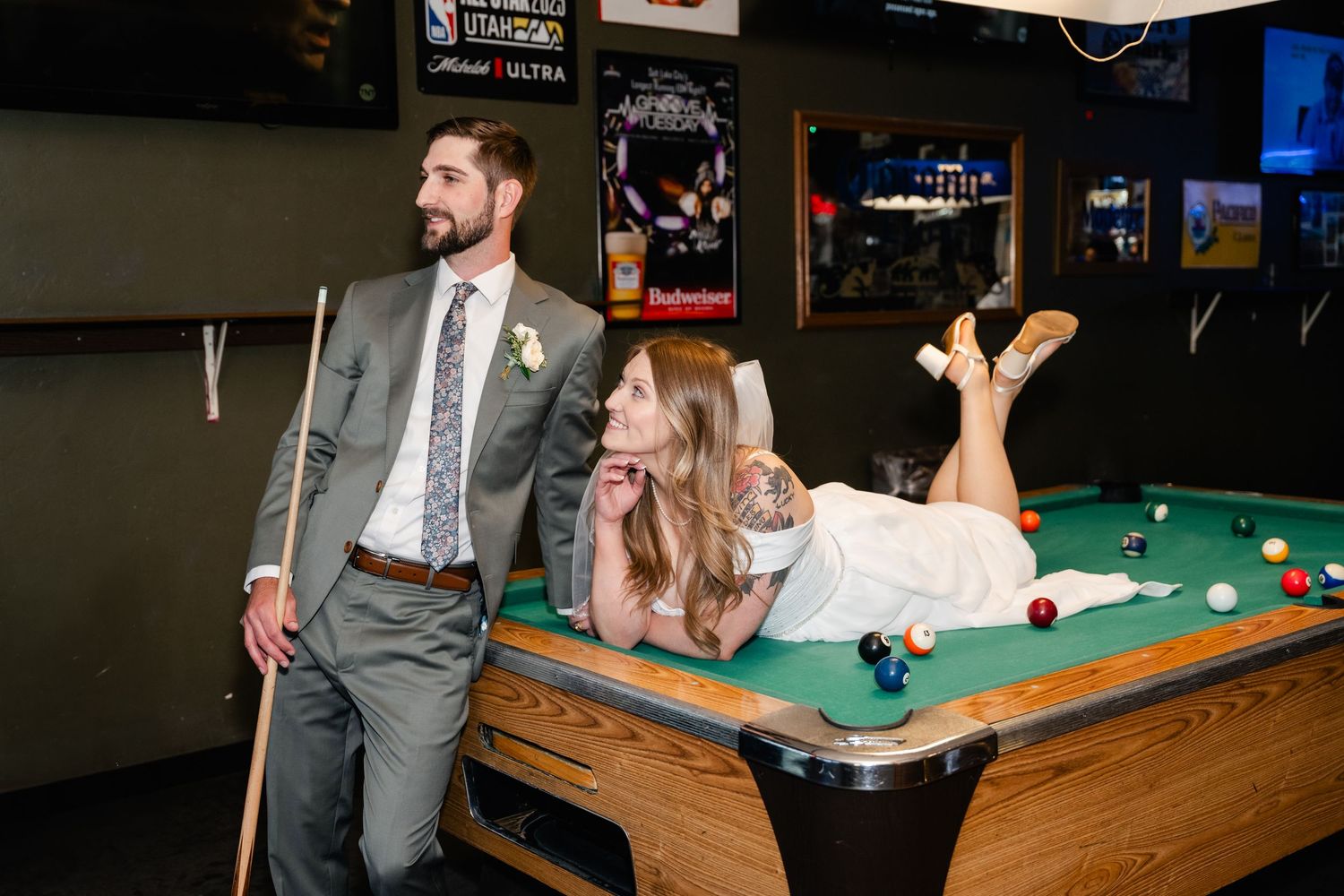 A couple poses playfully at a pool table in a bar setting with the bride lying on the table while wearing her wedding dress.