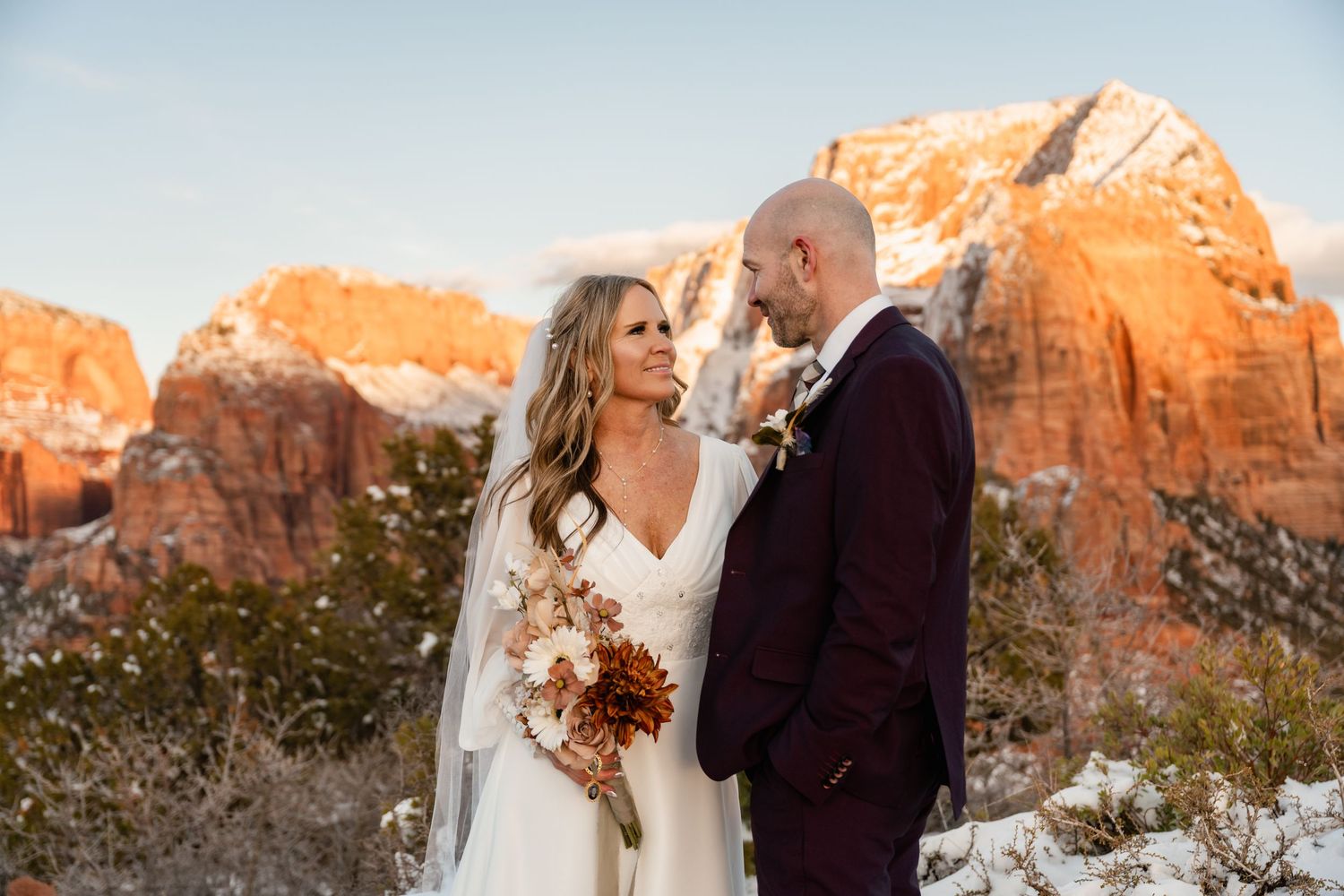 A romantic winter wedding photo shoot against stunning red rock cliffs and snowy landscape at sunset.