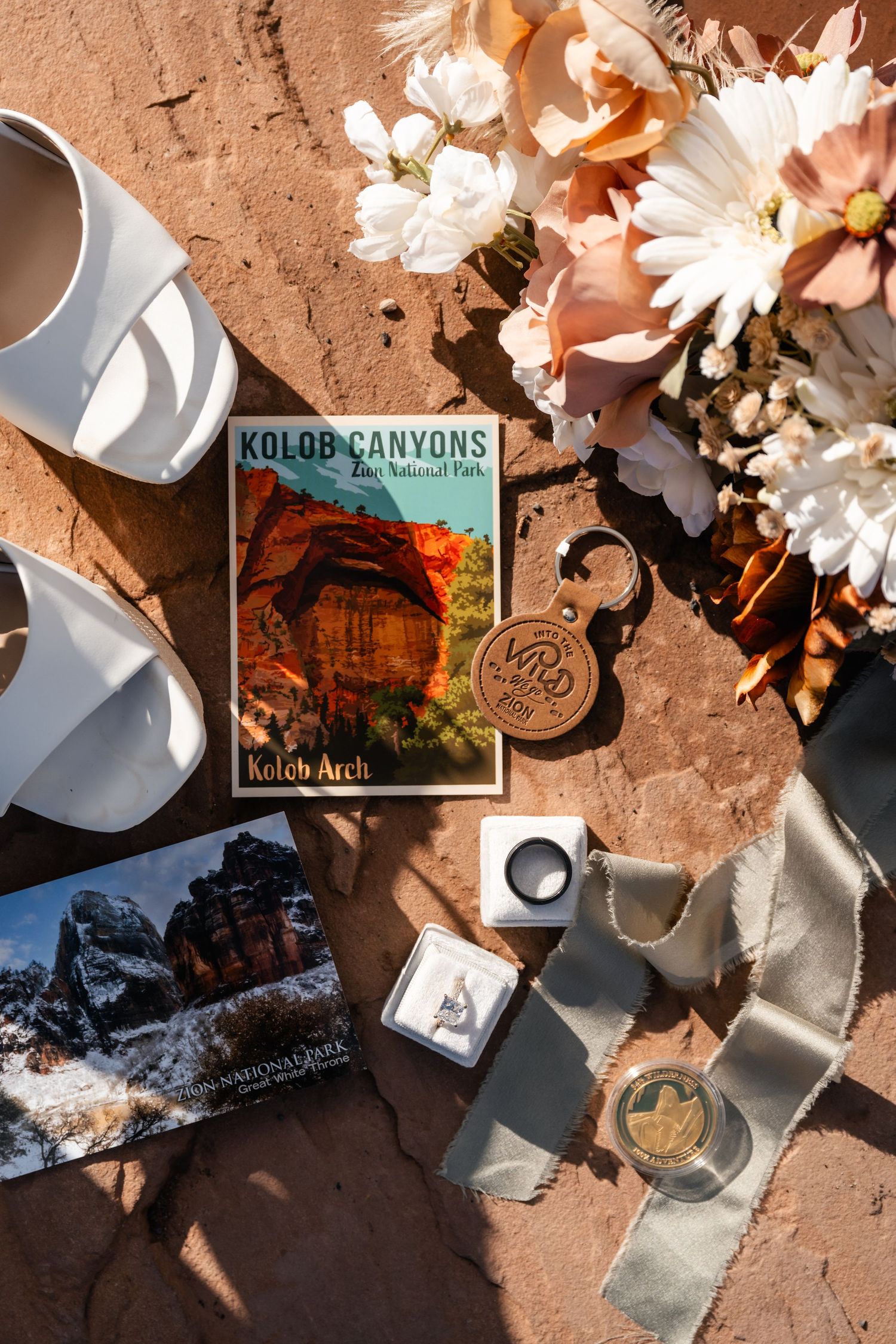 Flatlay of travel items including flowers, coffee cup, and Kolob Canyons postcard on red sandstone surface.