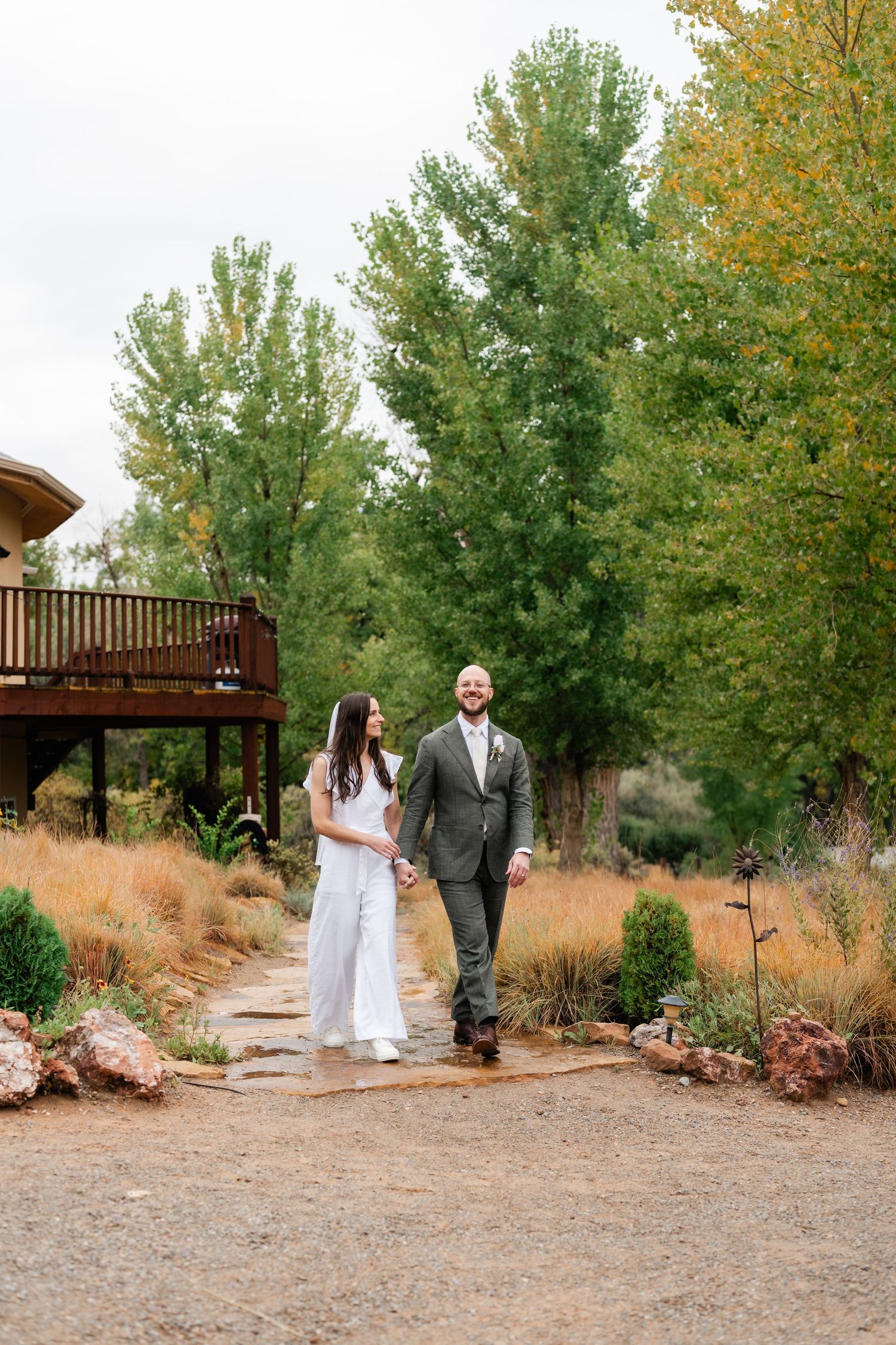 A couple in wedding attire walks along a gravel path lined with autumn trees.