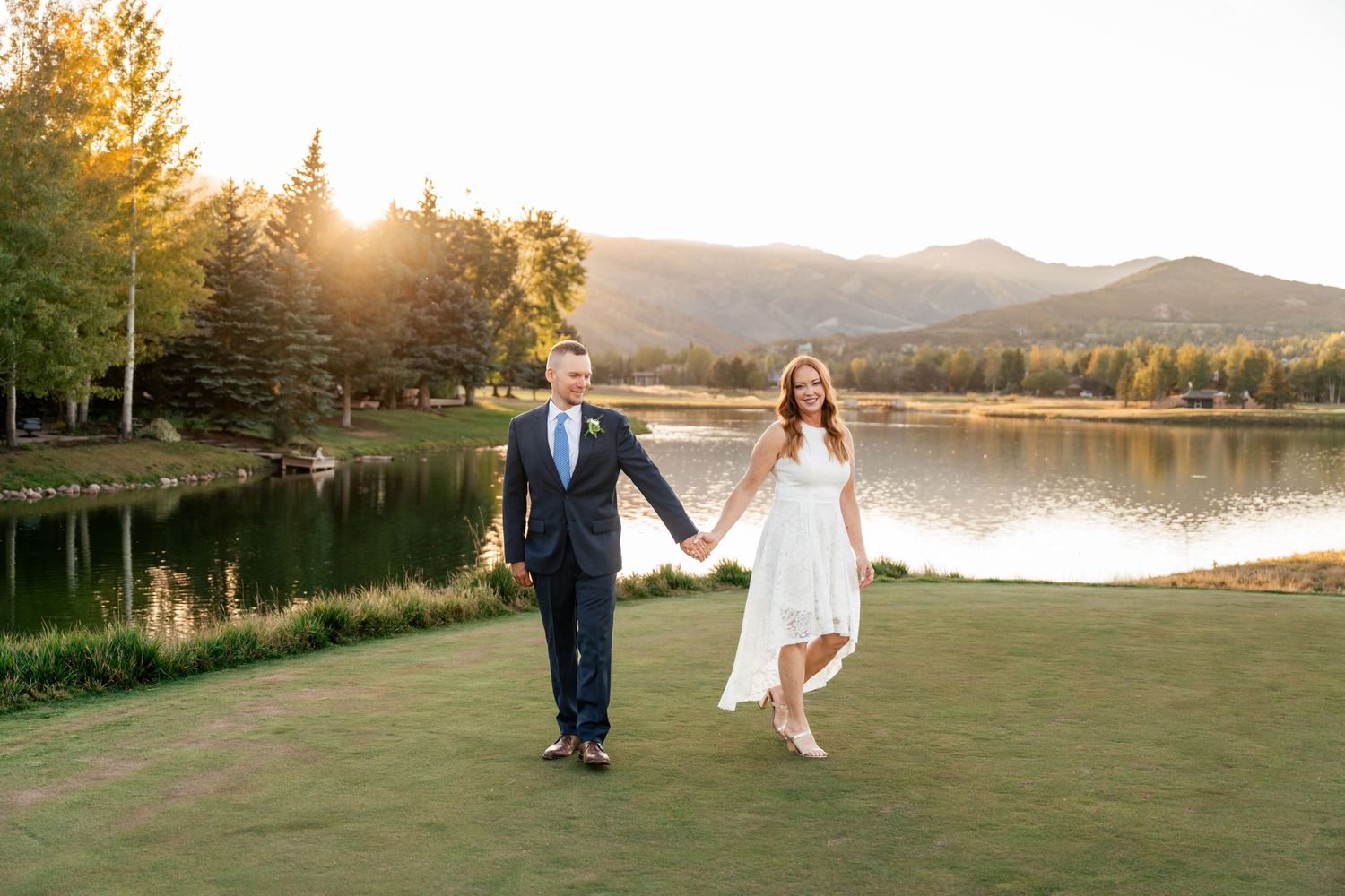 A couple walks along a scenic lakeside path during golden hour with mountains and autumn trees in the background.