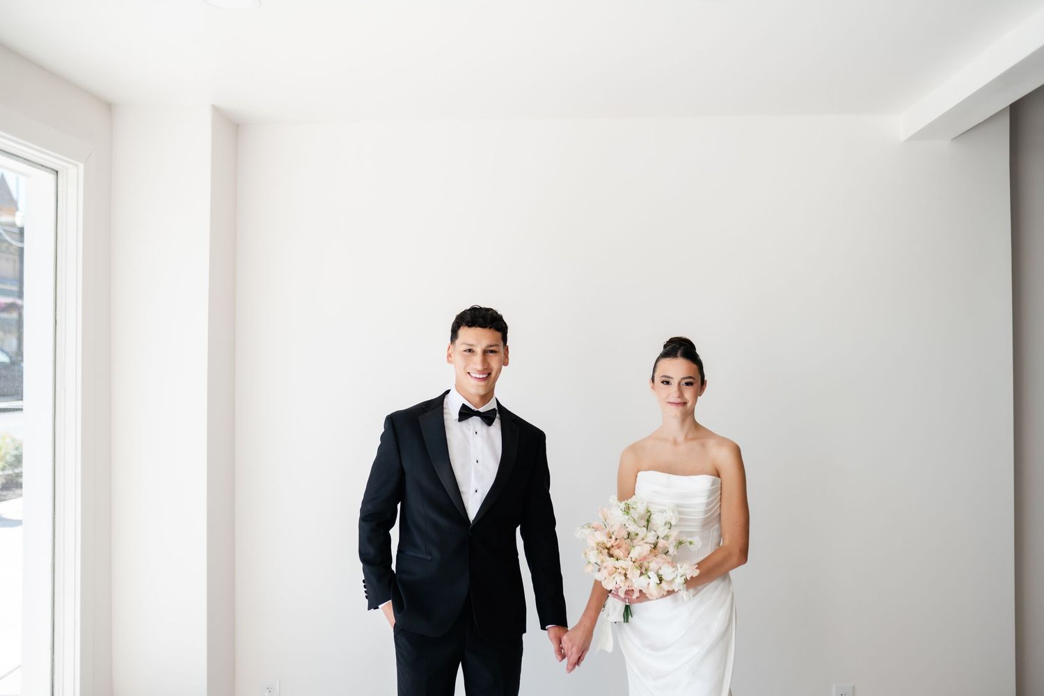 A bride and groom pose together in a minimalist white room, with the bride holding a soft neutral bouquet. Moments on Main Venue.