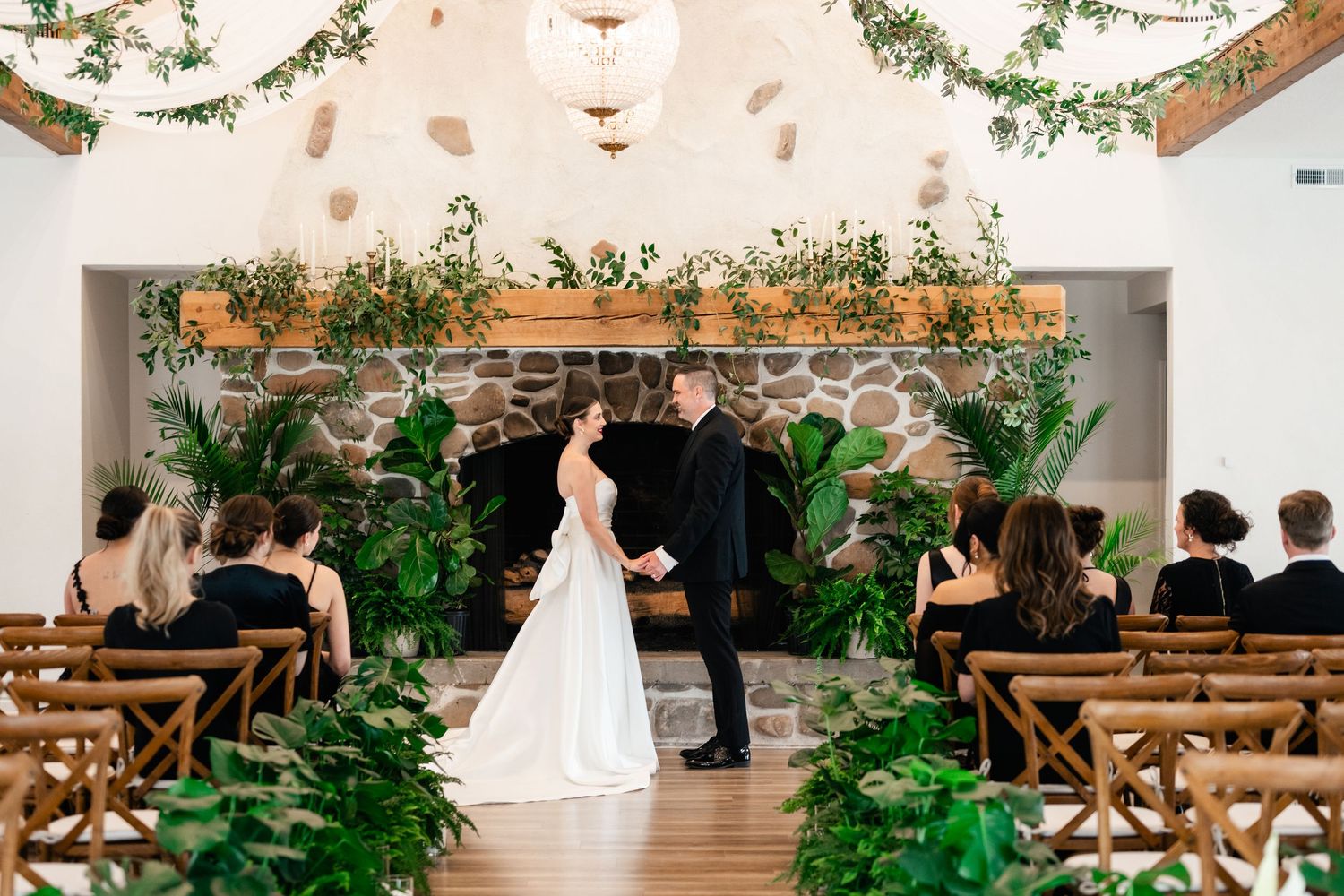 A rustic indoor wedding ceremony venue with wooden benches, white walls, and lush green plants decorating the space.