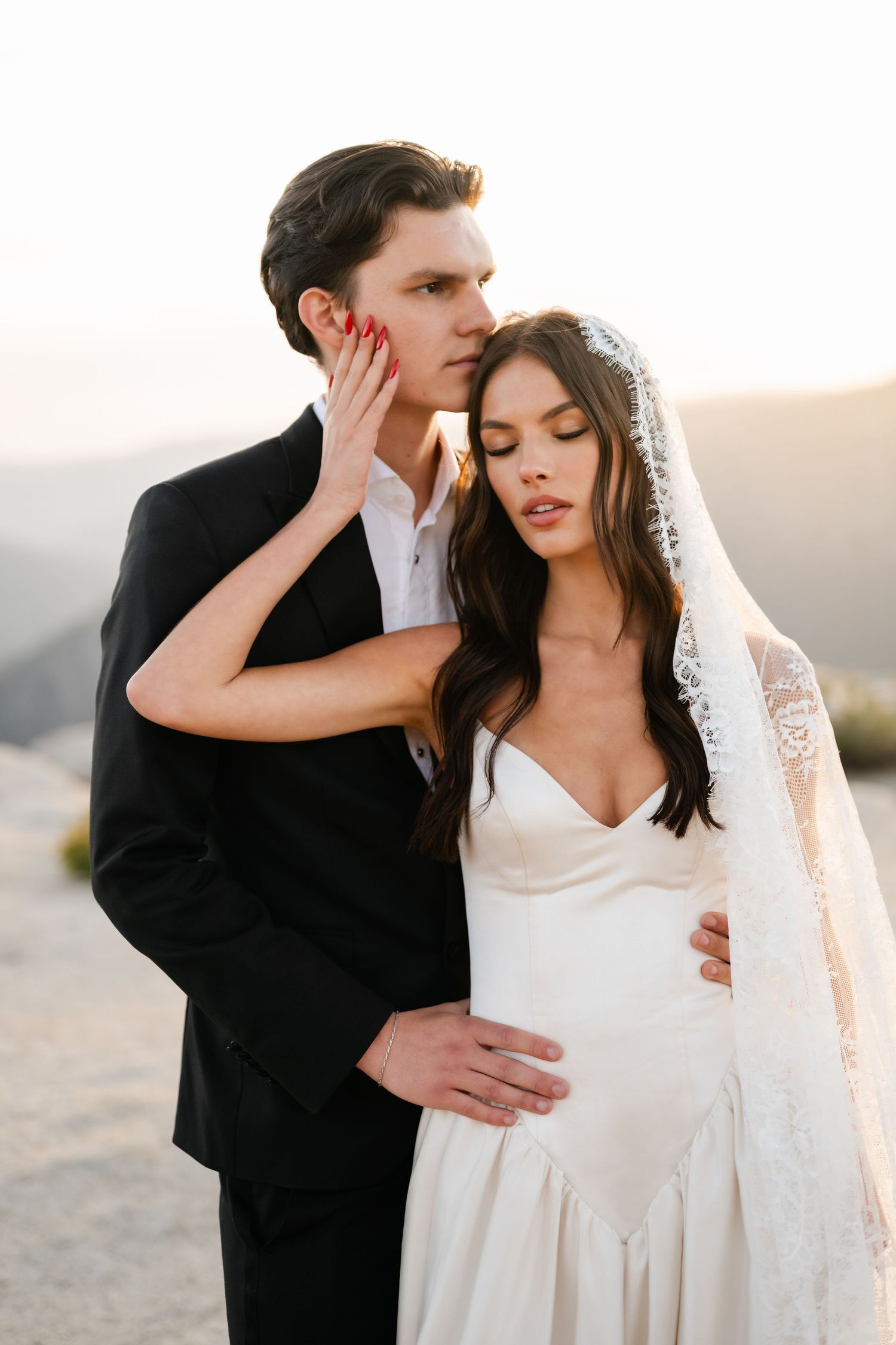 Intimate close-up of bride in silk wedding gown touching grooms face at golden hour mountain backdrop.