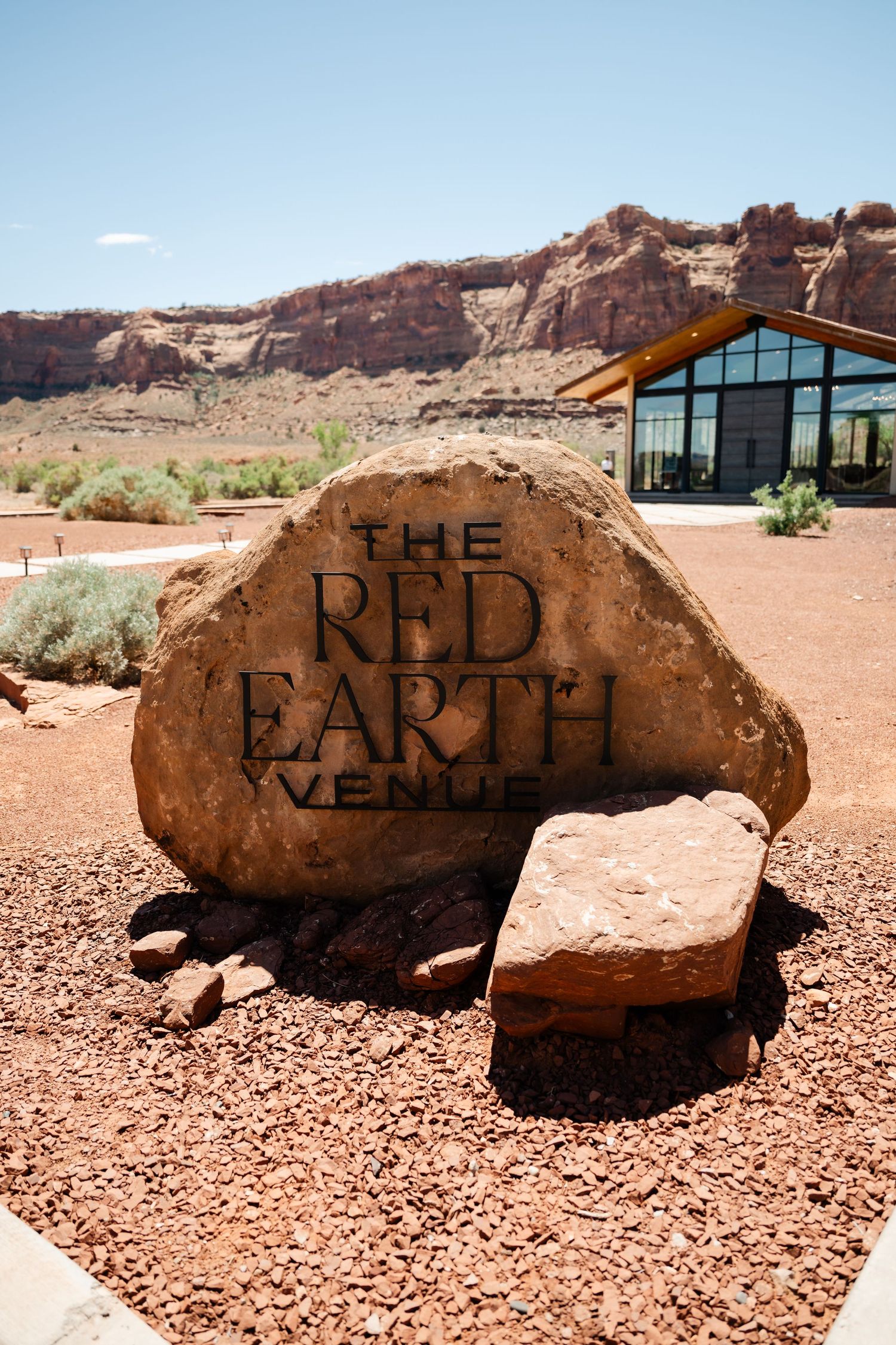The Red Earth Venue sign carved into a large sandstone boulder in a desert landscape with red rock formations.