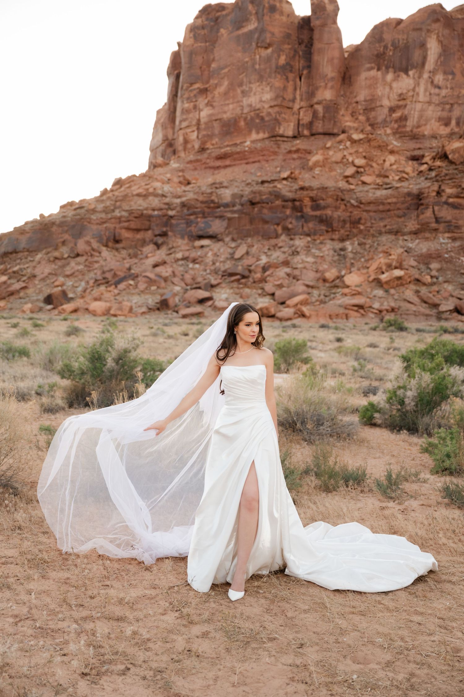 Bride in strapless white gown with flowing veil stands before red rock formations in desert.
