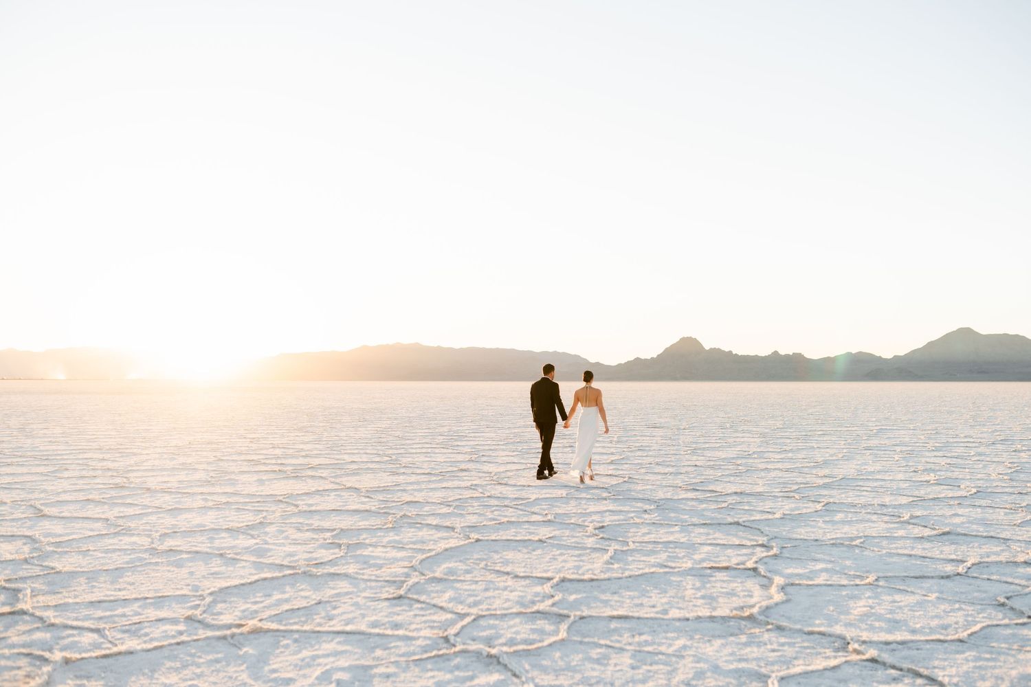 Two silhouettes walk hand in hand across the vast white salt flats at sunset with mountains in the distance. Salt Flats Photographer.