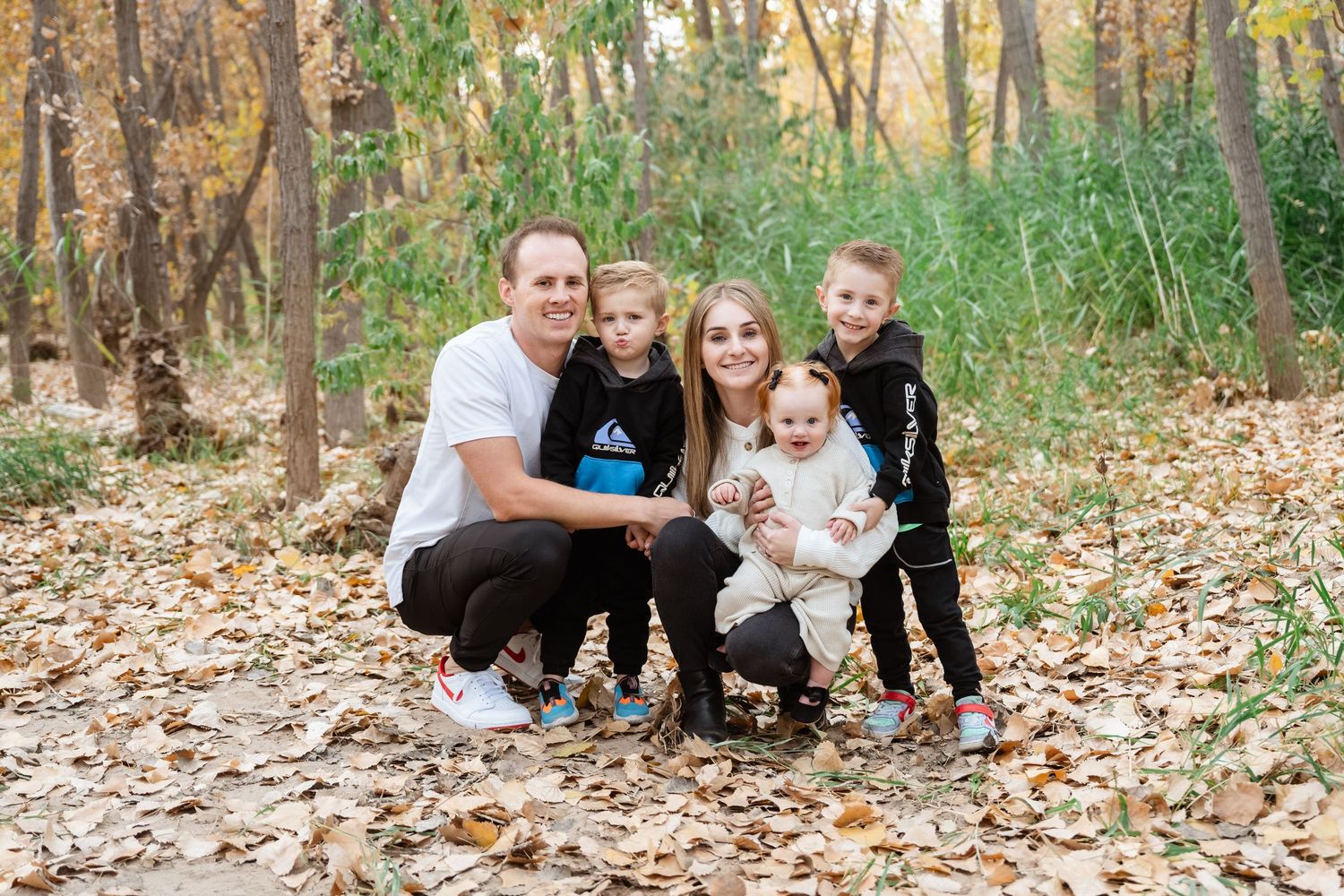 A family sits together on fallen autumn leaves in a wooded area during a casual outdoor photo session.