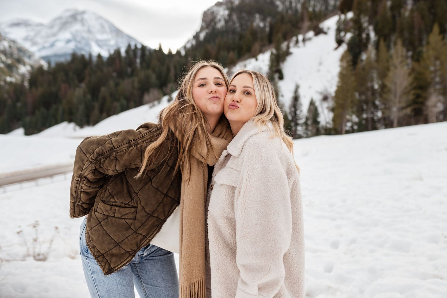 Two people embrace while standing in a snowy winter landscape with snow-covered mountains and evergreen trees.