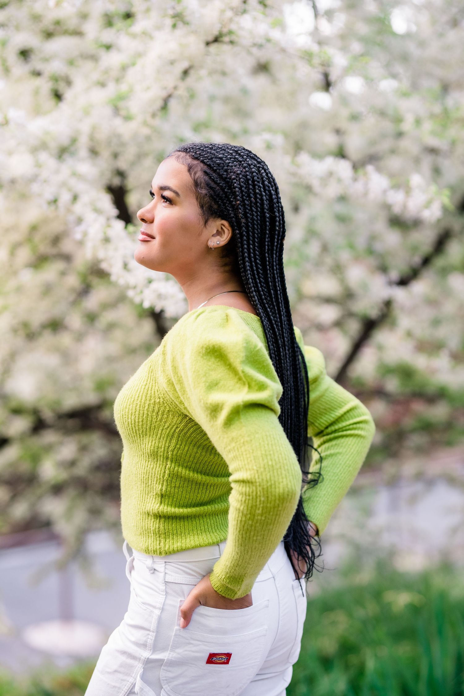 Person in bright lime green sweater and white pants poses gracefully against blooming cherry blossom trees in spring.