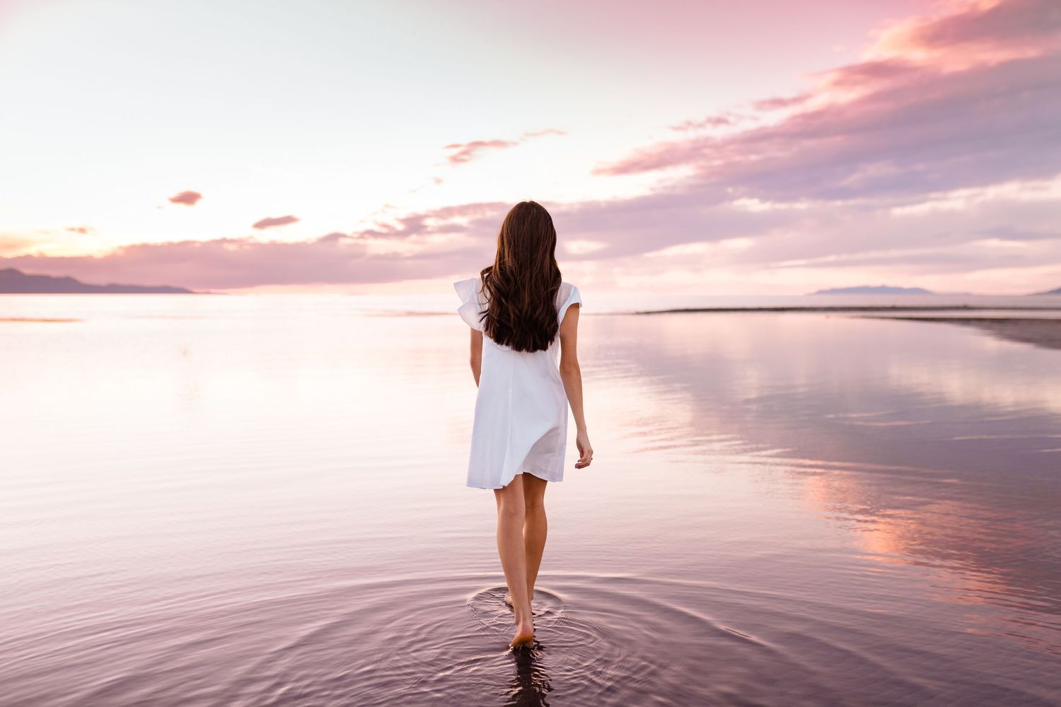 A person walks along a tranquil beach at sunset with pink and purple clouds reflecting in the calm ocean water.