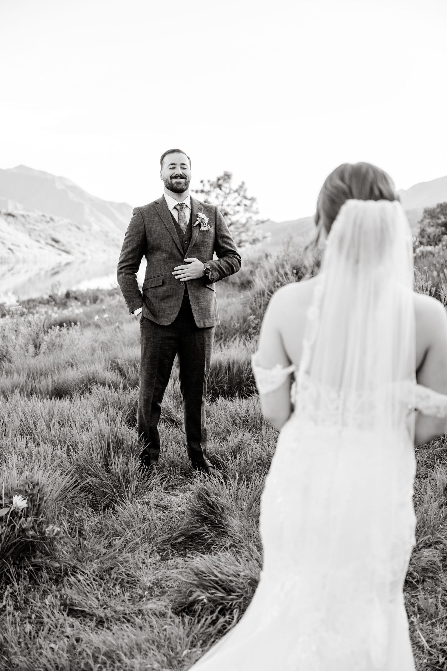 Black and white wedding portrait showing a bride in flowing dress approaching groom on a grassy mountainside.