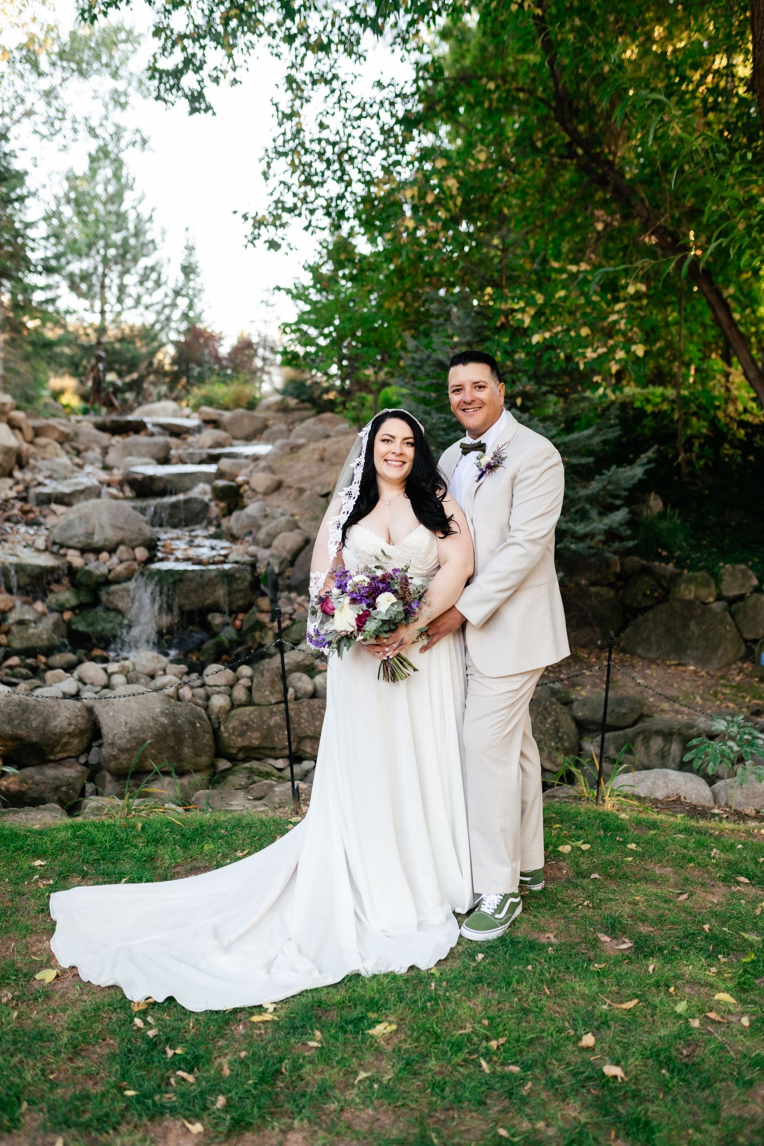 A couple in wedding attire poses by a waterfall feature with the bride holding a purple bouquet.