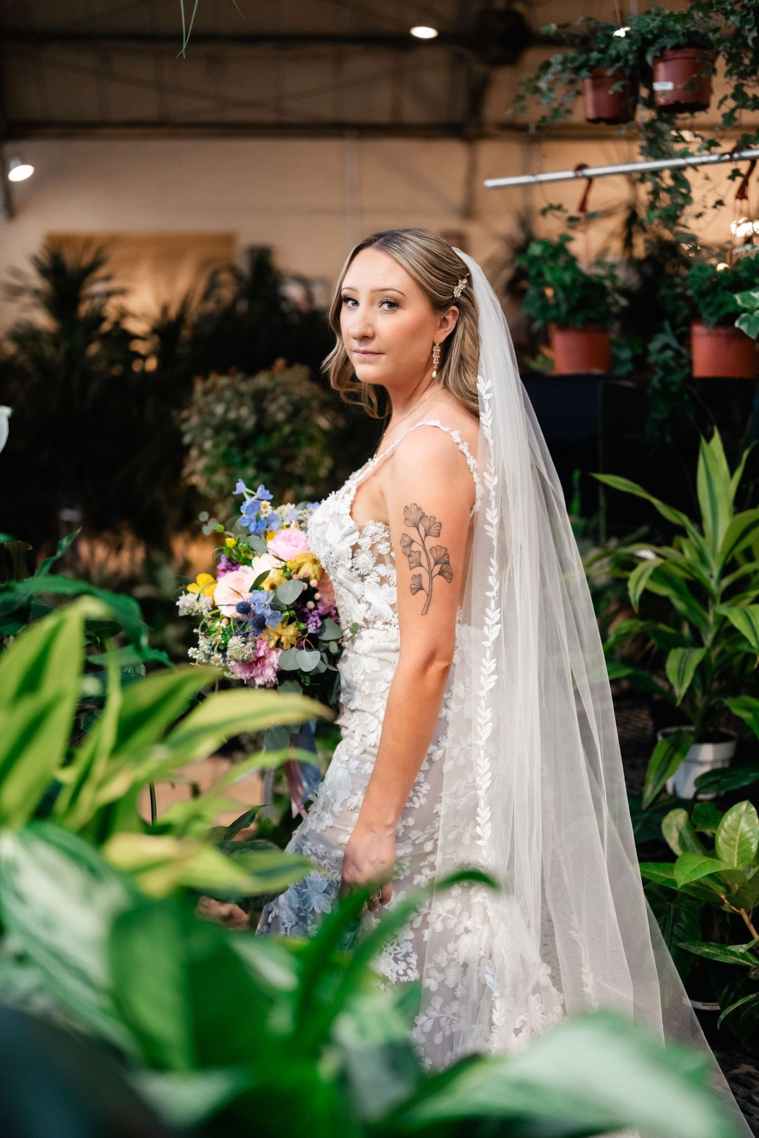 Elegant bride in floral detailed dress holds colorful bouquet while standing among lush indoor greenery and potted plants.