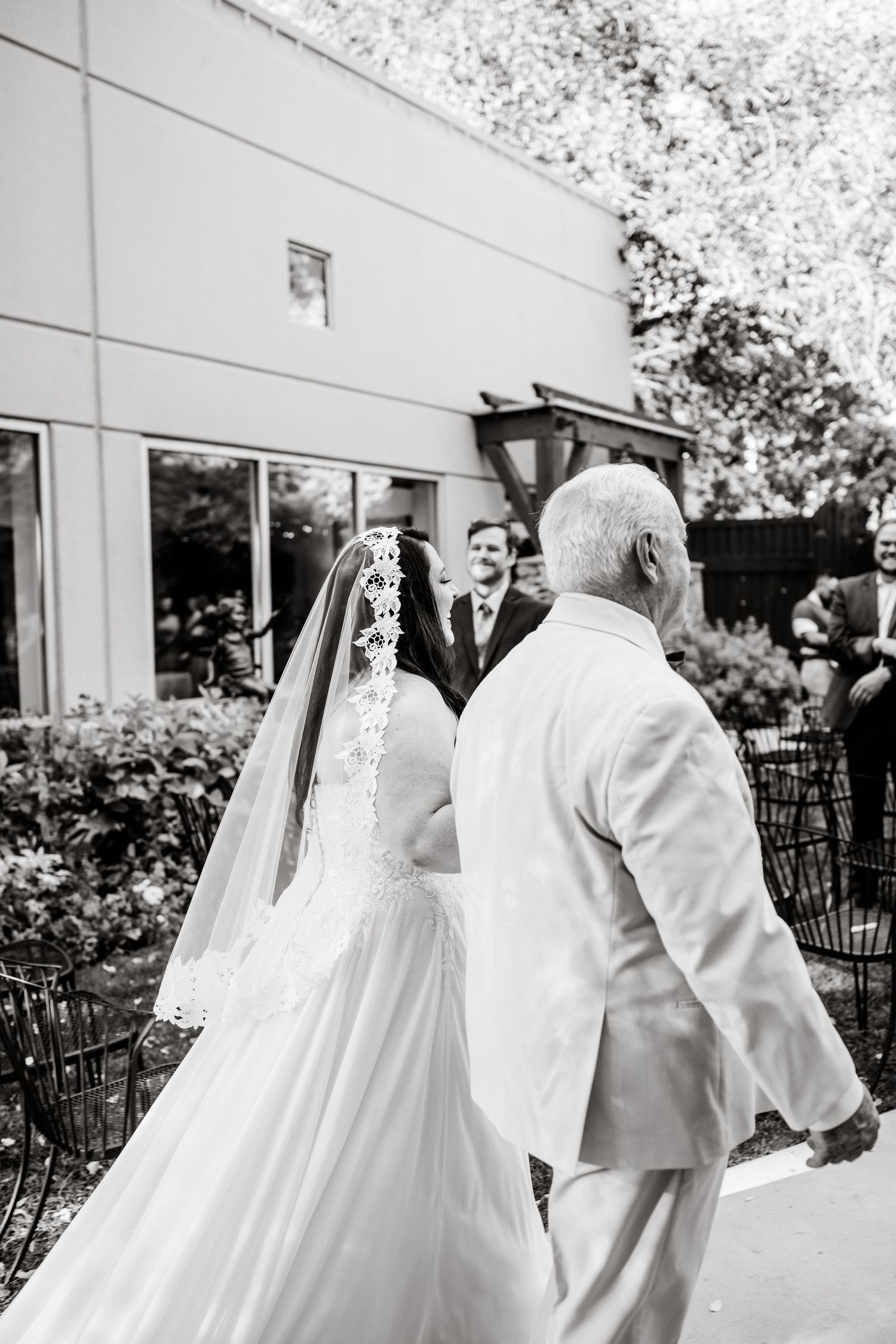 A bride in a flowing white dress and veil walks alongside a guest in a light colored suit during an outdoor wedding.