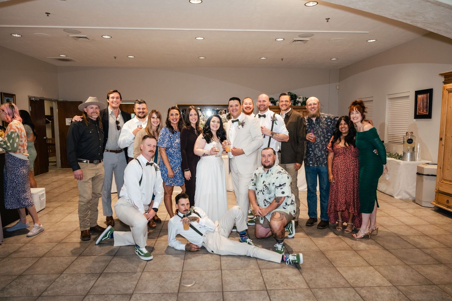 Large group poses for a casual wedding photo on a tiled floor with white walls and decor.