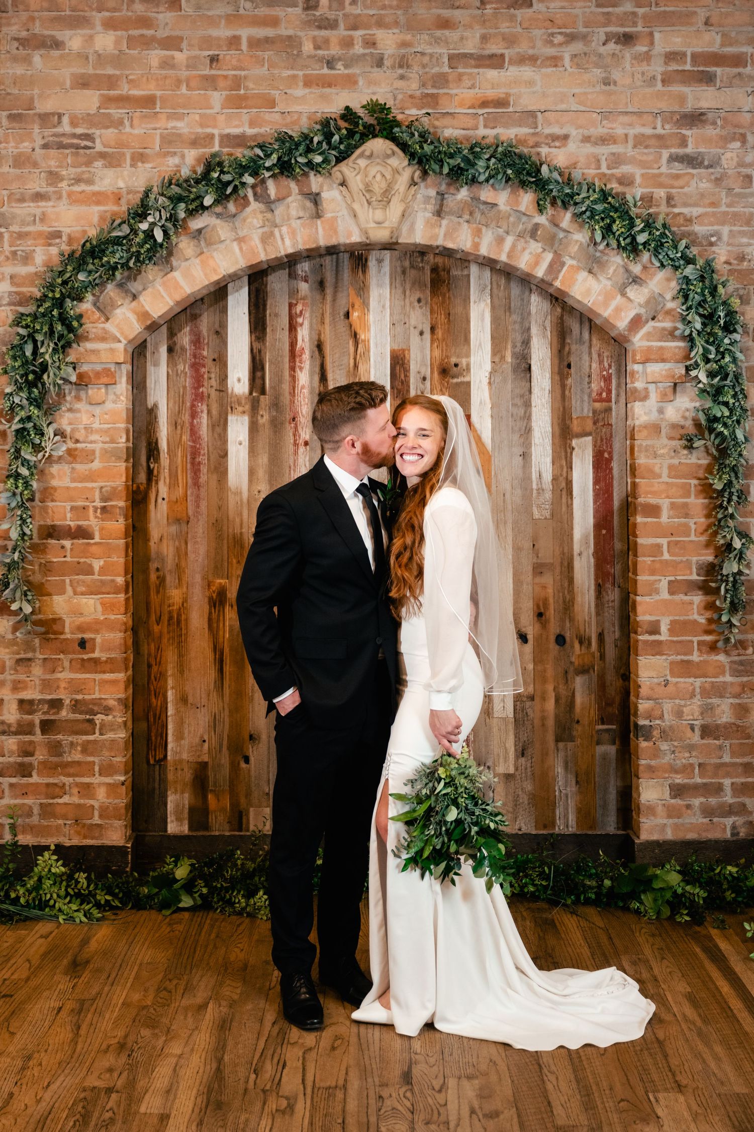 A romantic winter wedding scene featuring a rustic wooden door decorated with greenery garland at a brick venue.