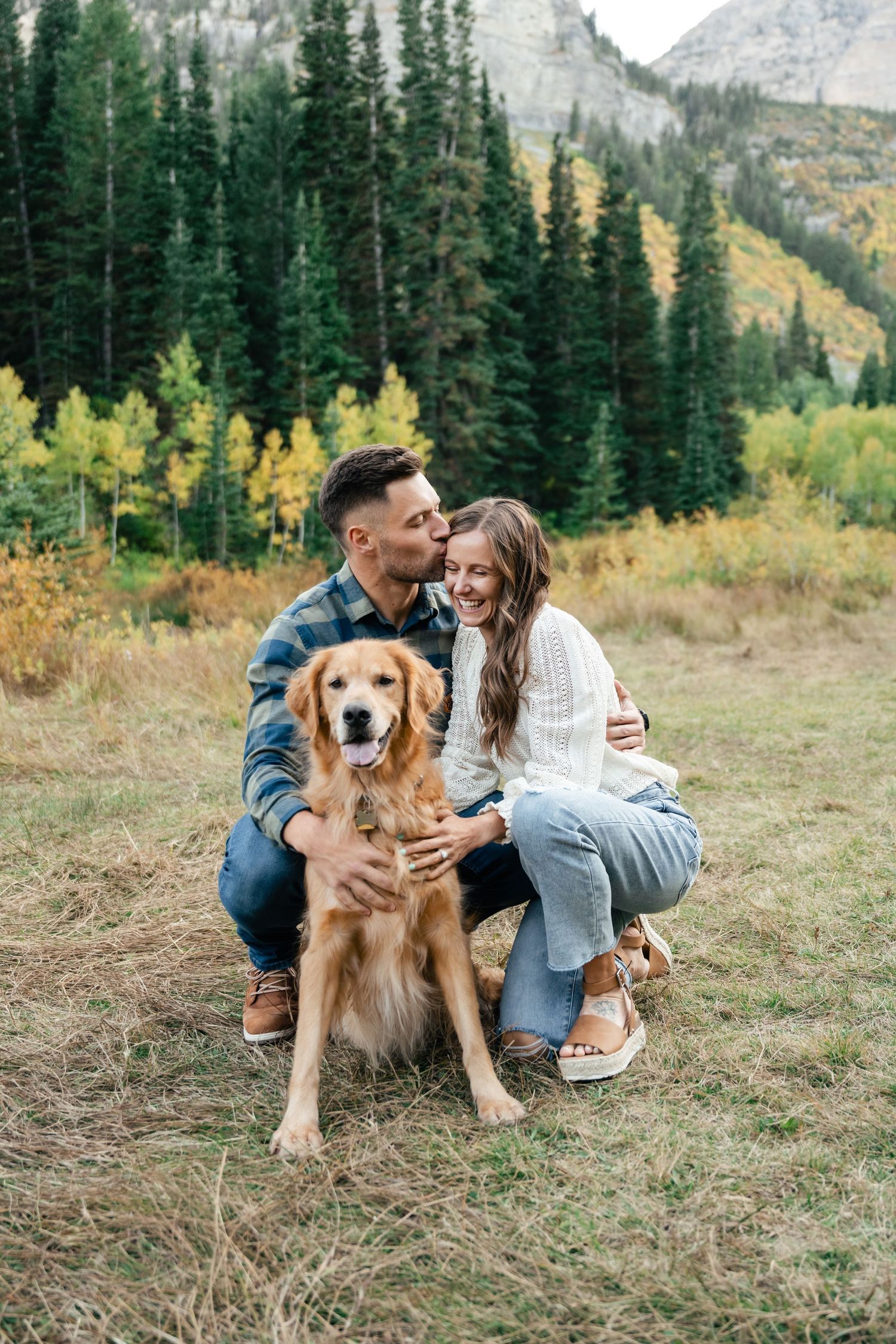 A happy couple poses with their Golden Retriever dog against a backdrop of mountain scenery and fall colors.