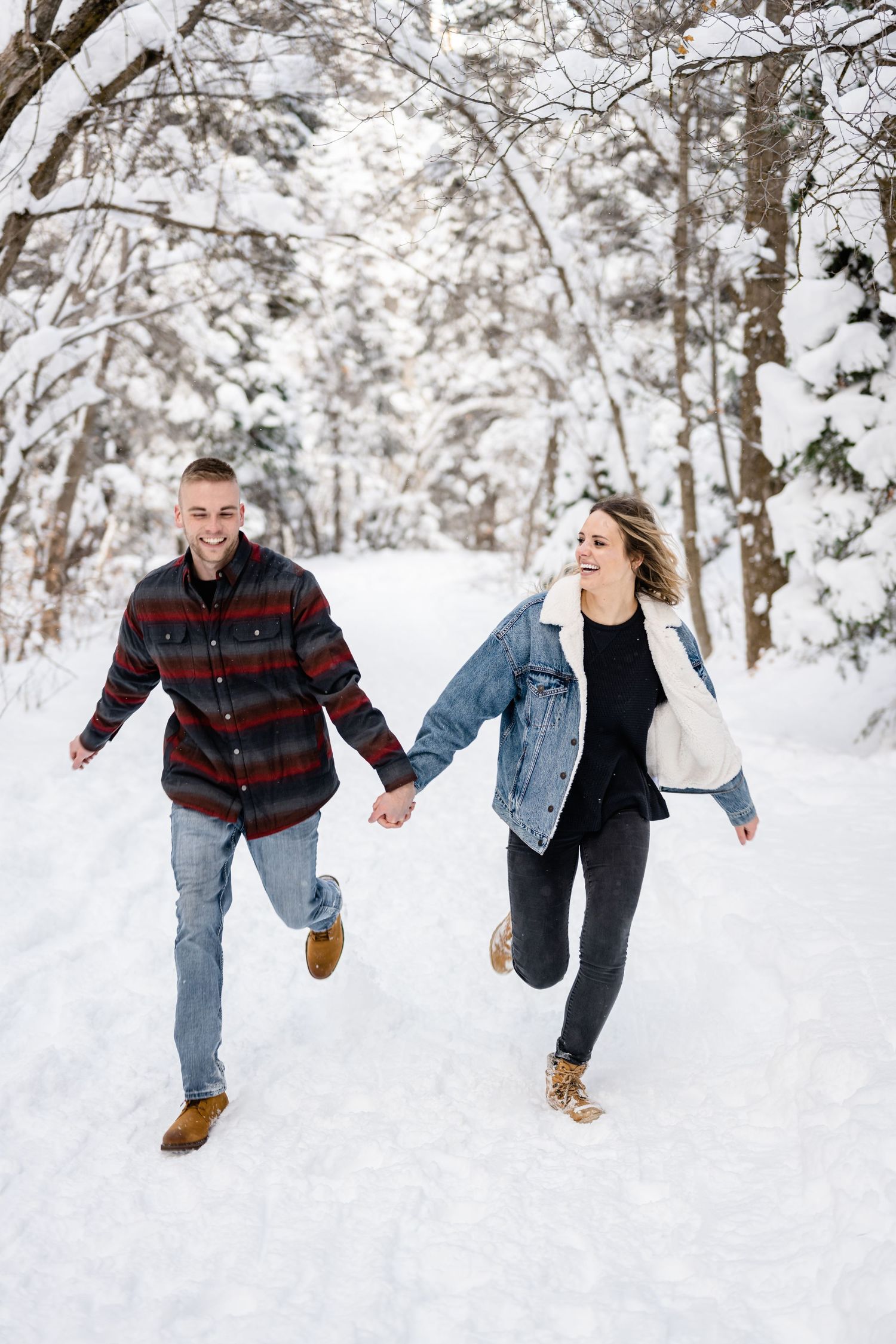 A couple runs joyfully hand-in-hand through a snowy forest path wearing winter clothing.