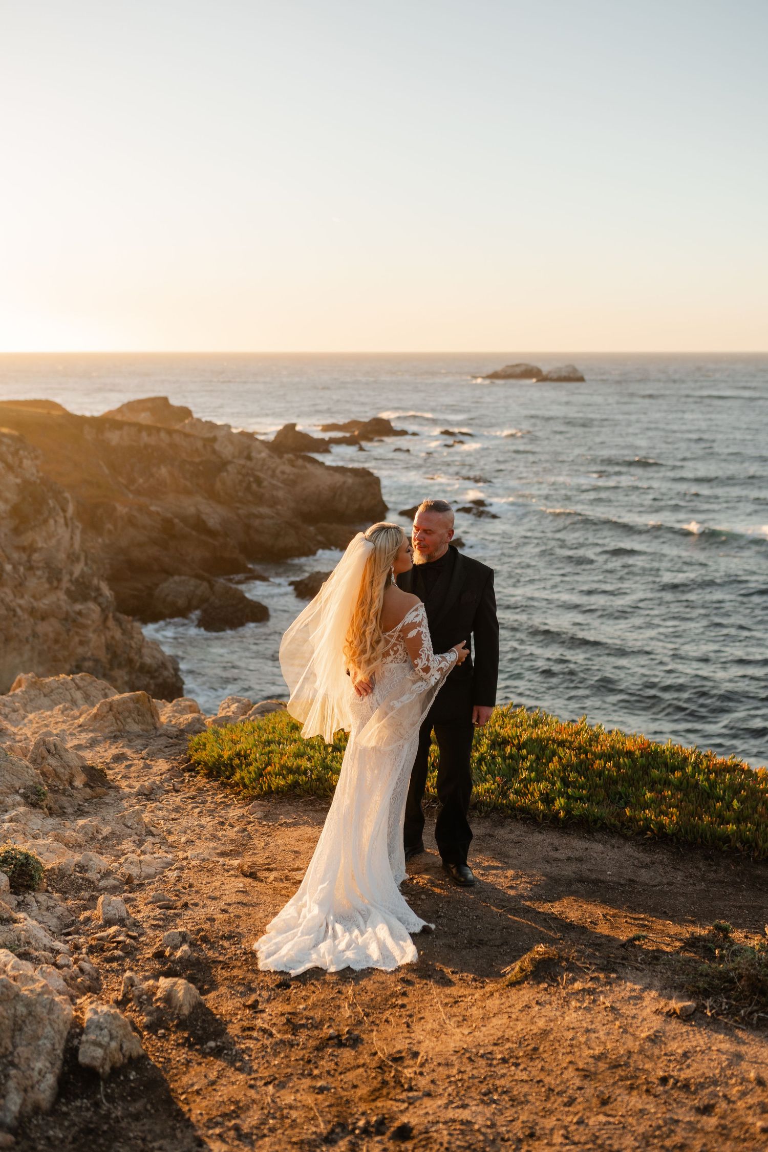 A romantic coastal wedding portrait at sunset with rocky cliffs and ocean waves.