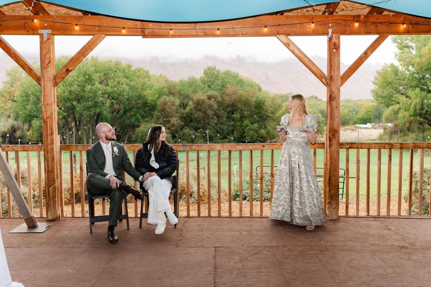 Wedding party members seated along wooden railing of rustic outdoor pavilion.
