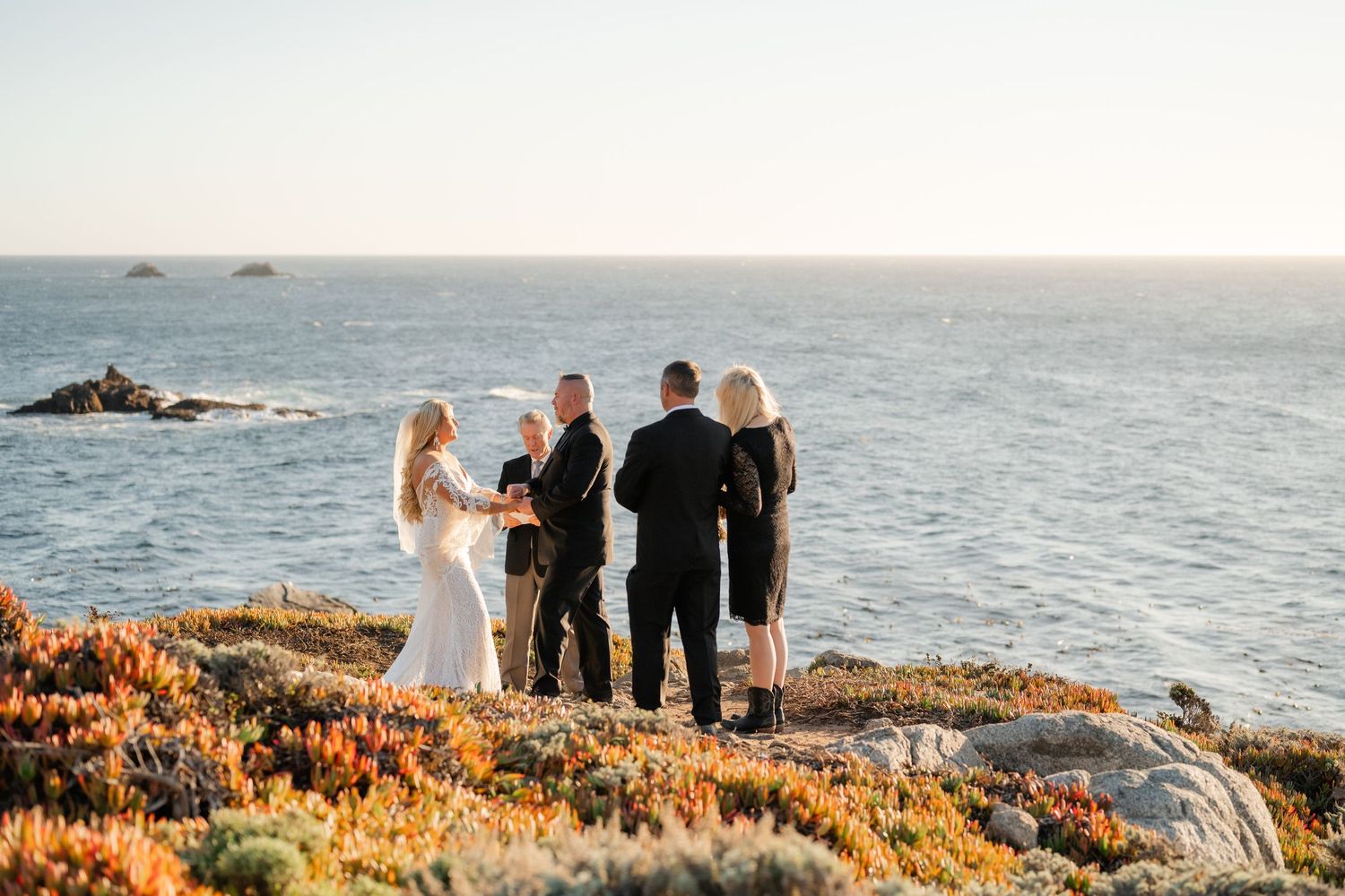 Panoramic view of a beach wedding ceremony with guests gathered on the rocky coastline at sunset.